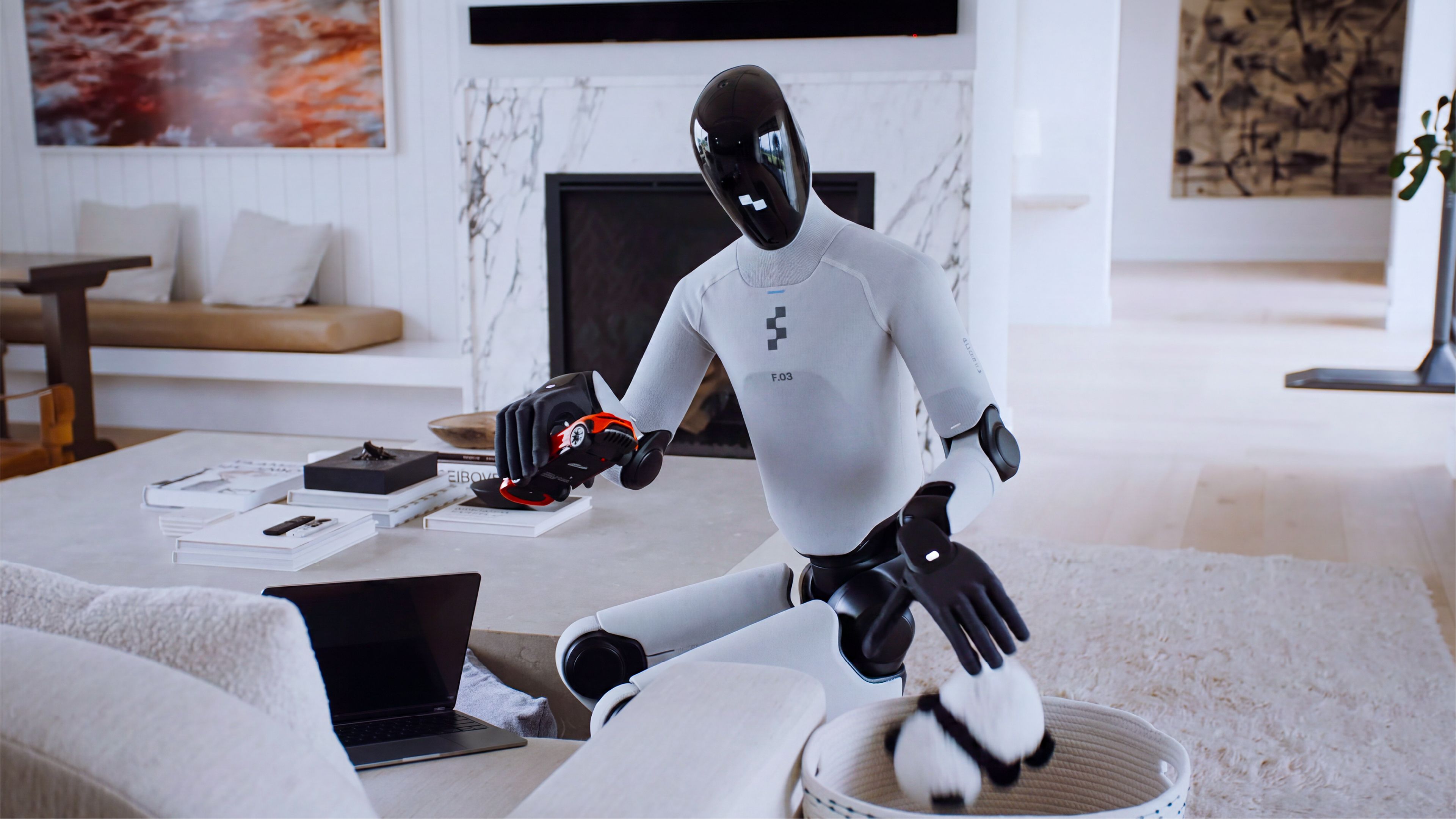A humanoid robot with a glossy black helmet-shaped head and white-and-black body, labeled "F.03," sits at a coffee table in a bright, modern living room. It holds a small toy car in one hand while reaching toward a stuffed toy in a woven basket with its other hand. A laptop, books, and decorative objects sit on the table nearby, with a marble fireplace, framed artwork, and soft furnishings visible in the background.