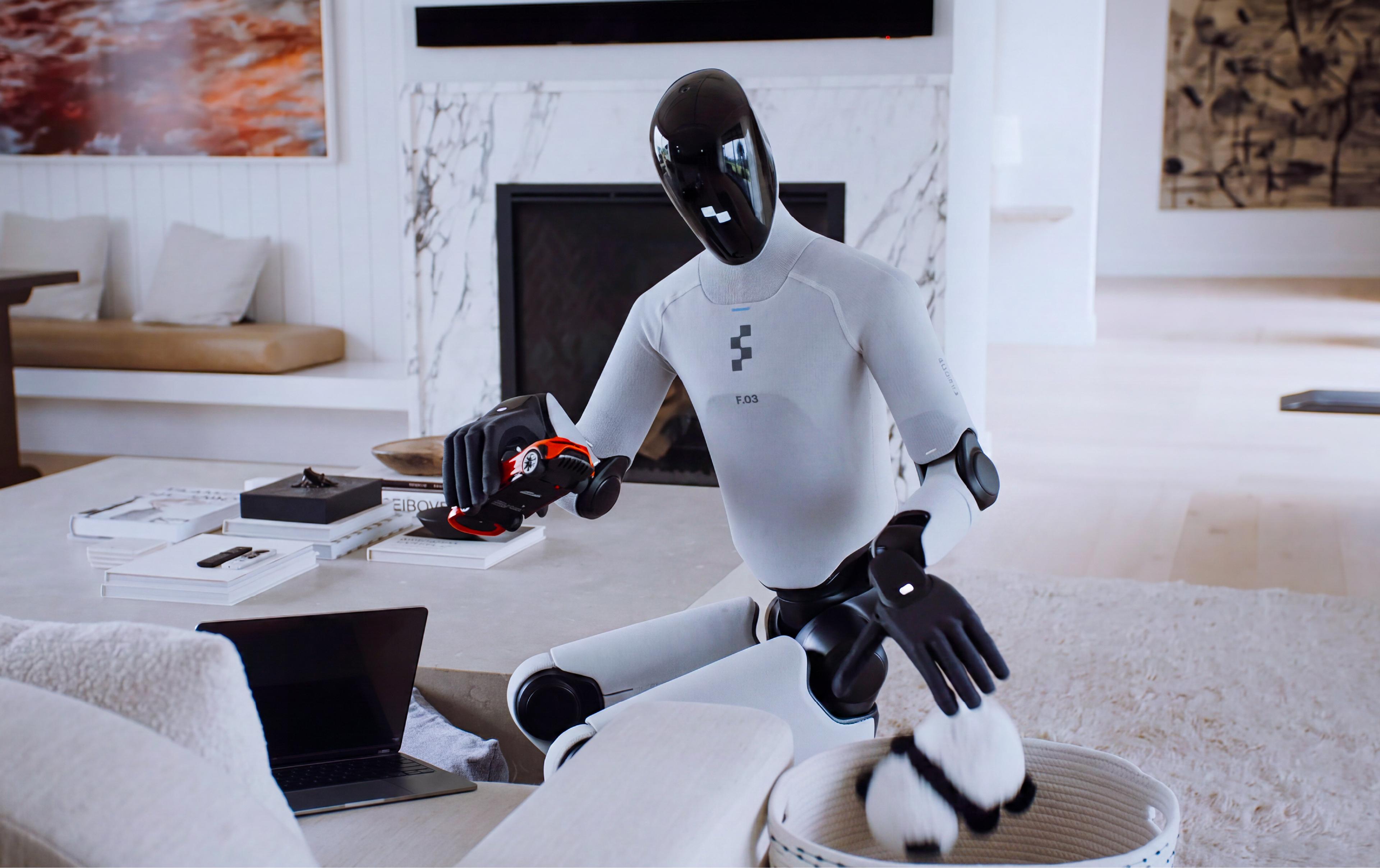 A humanoid robot with a glossy black helmet-shaped head and white-and-black body, labeled "F.03," sits at a coffee table in a bright, modern living room. It holds a small toy car in one hand while reaching toward a stuffed toy in a woven basket with its other hand. A laptop, books, and decorative objects sit on the table nearby, with a marble fireplace, framed artwork, and soft furnishings visible in the background.