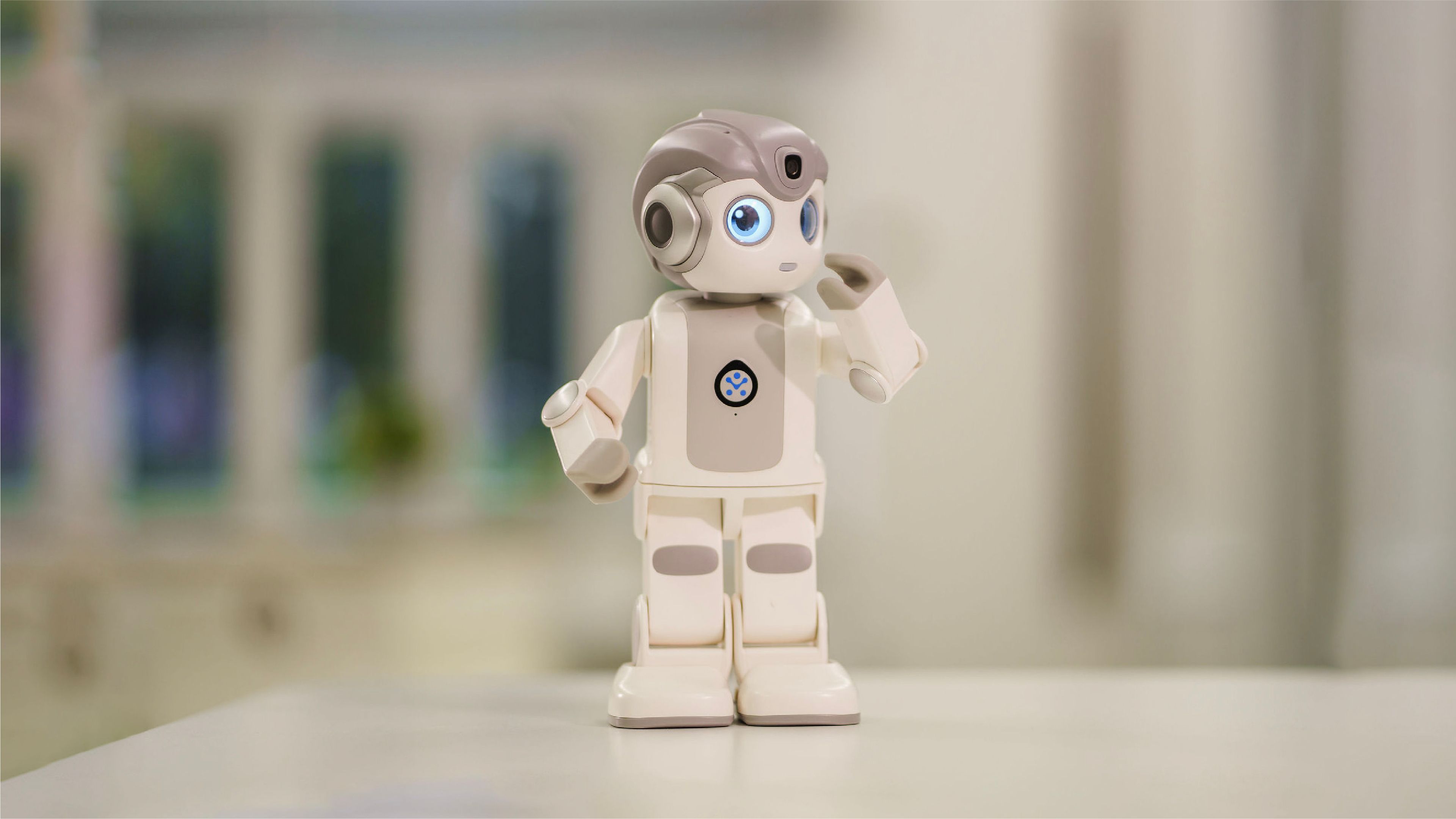 A robot stands on a table, with a window in the background allowing natural light to illuminate the scene.