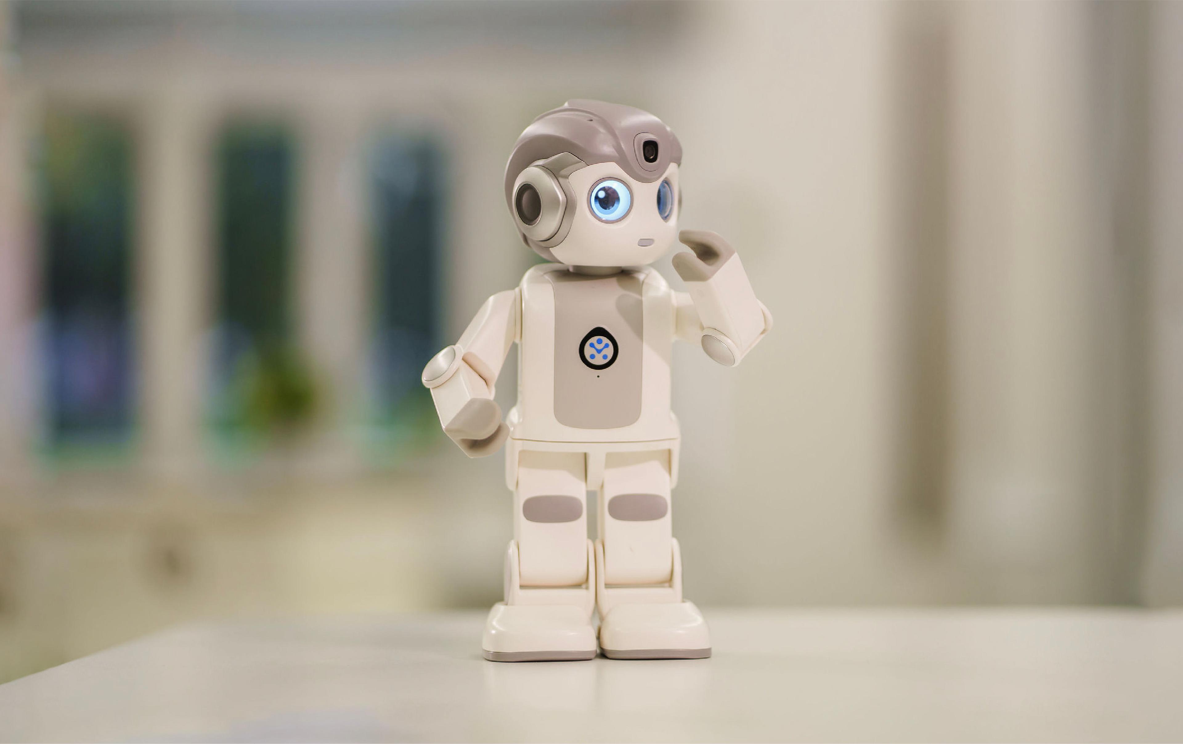 A robot stands on a table, with a window in the background allowing natural light to illuminate the scene.