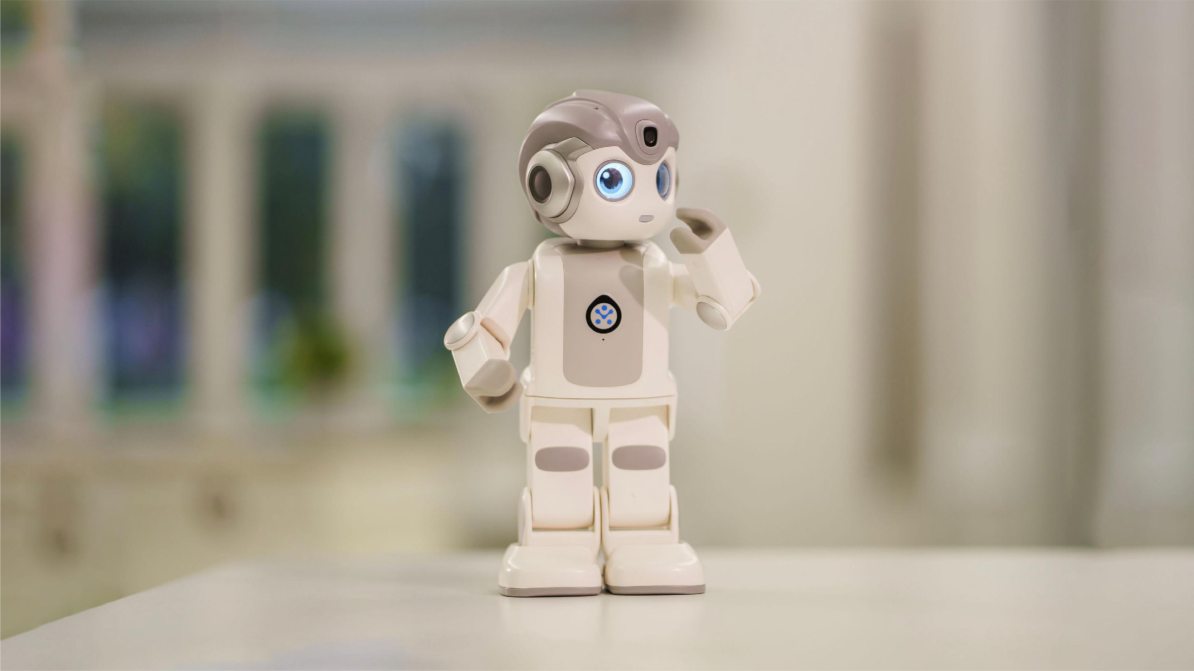 A robot stands on a table, with a window in the background allowing natural light to illuminate the scene.
