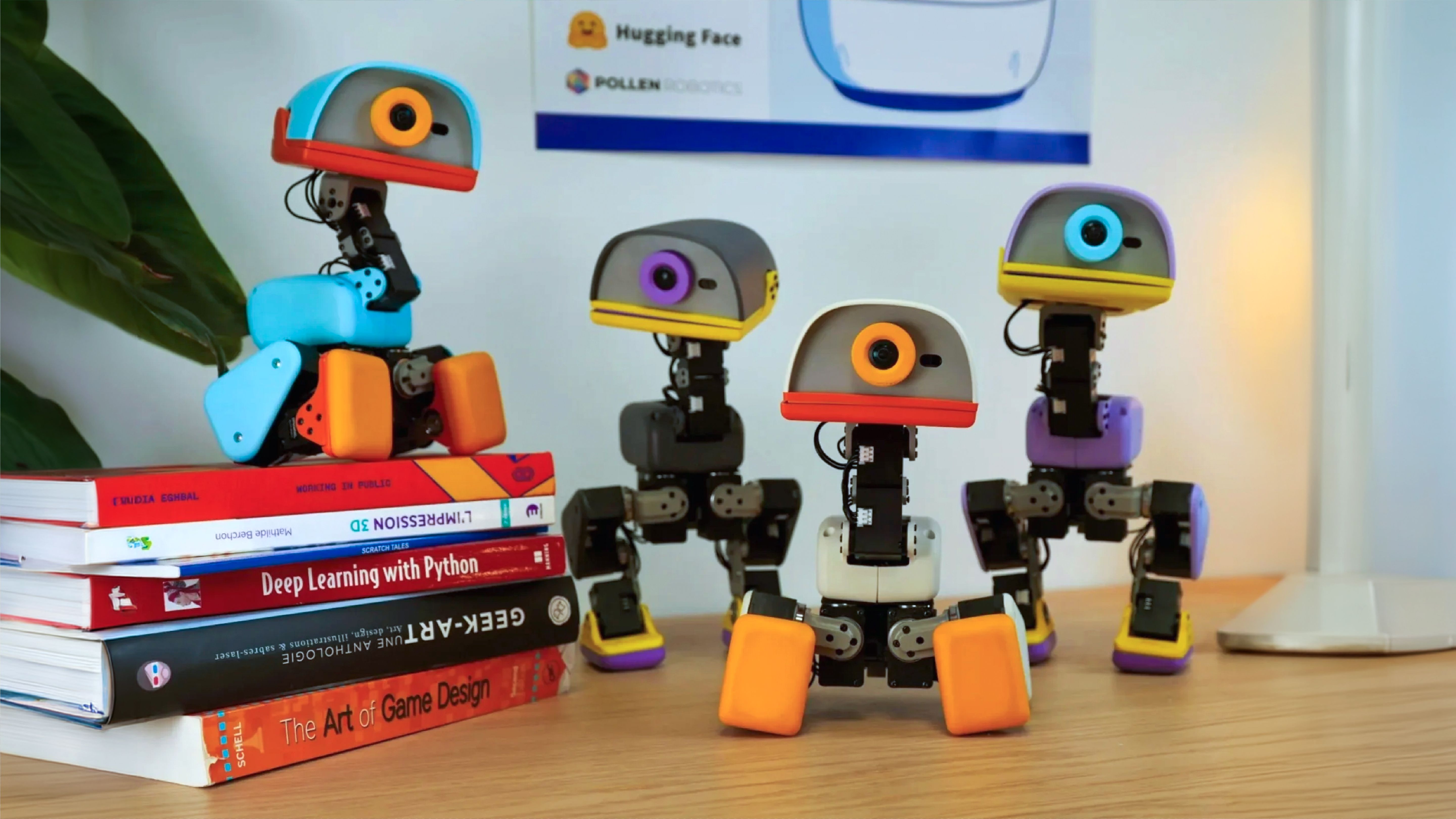 Four small bipedal duck robots stand on a desk beside a stack of books, with a Hugging Face and Pollen Robotics poster behind