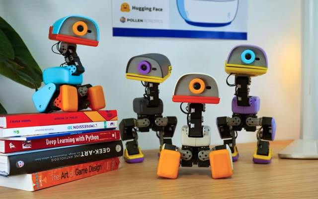 Four small bipedal duck robots stand on a desk beside a stack of books, with a Hugging Face and Pollen Robotics poster behind