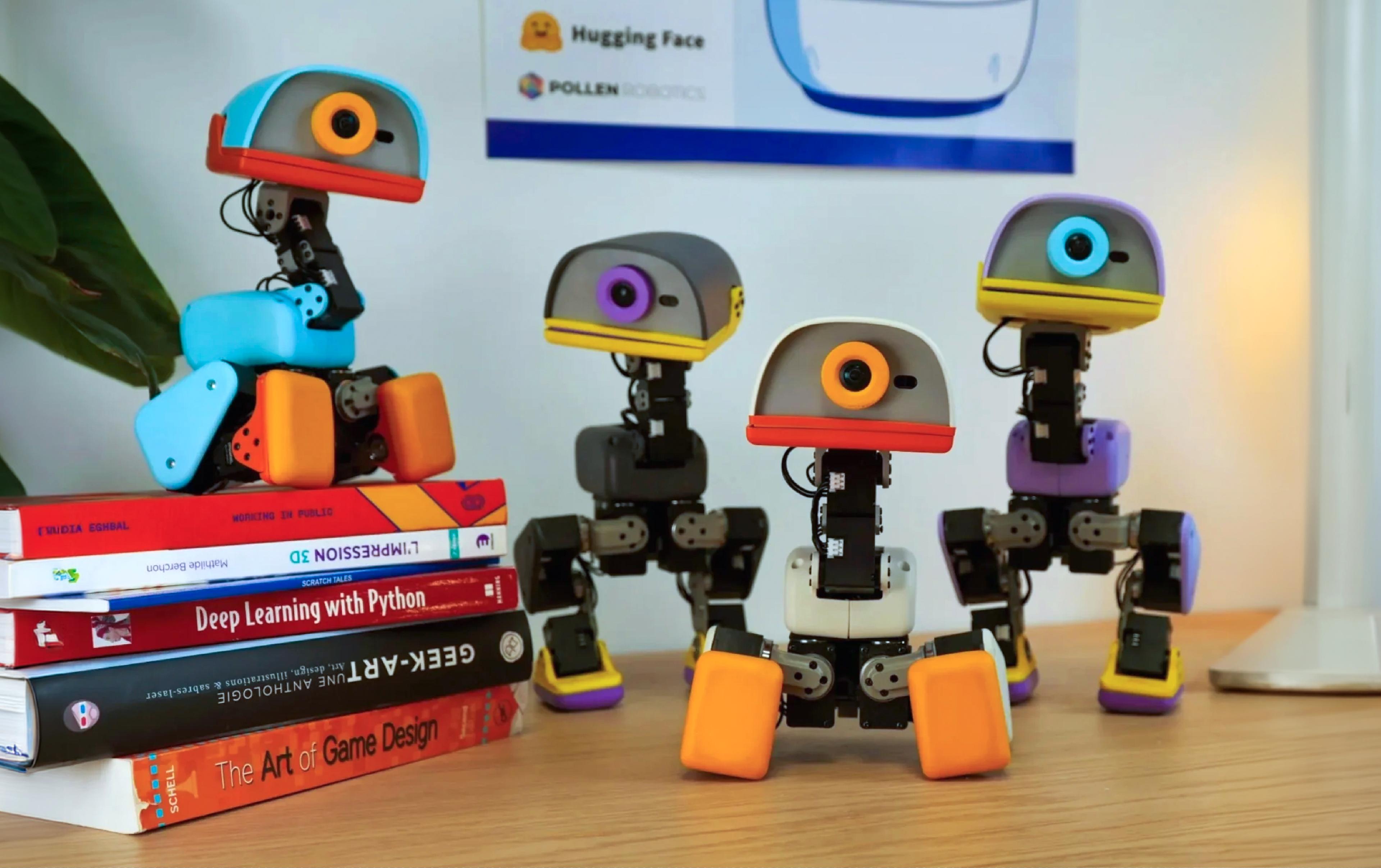 Four small bipedal duck robots stand on a desk beside a stack of books, with a Hugging Face and Pollen Robotics poster behind