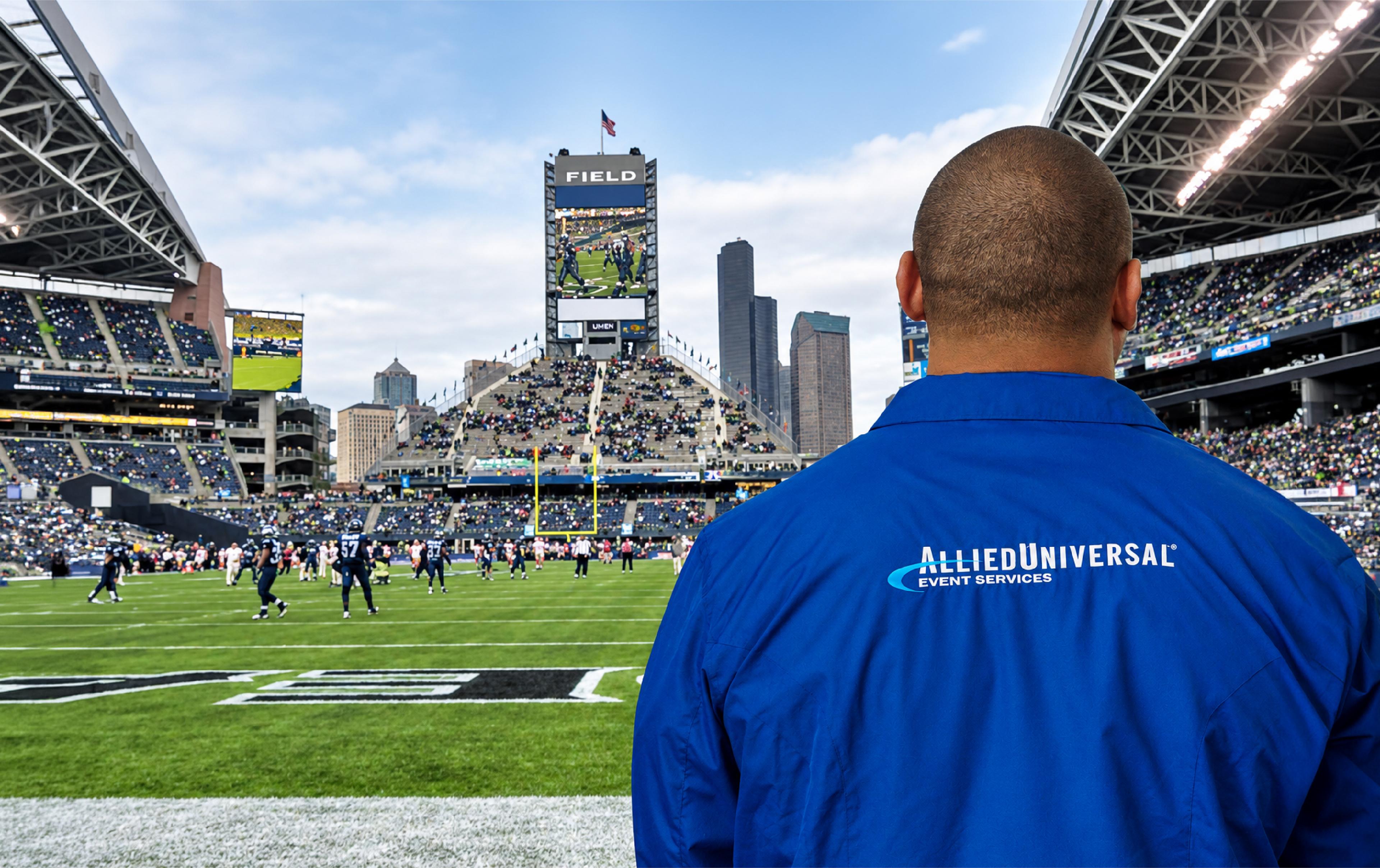 An Allied Universal Event Services employee overlooks the football field during a game at a packed stadium, with players and spectators in the background.