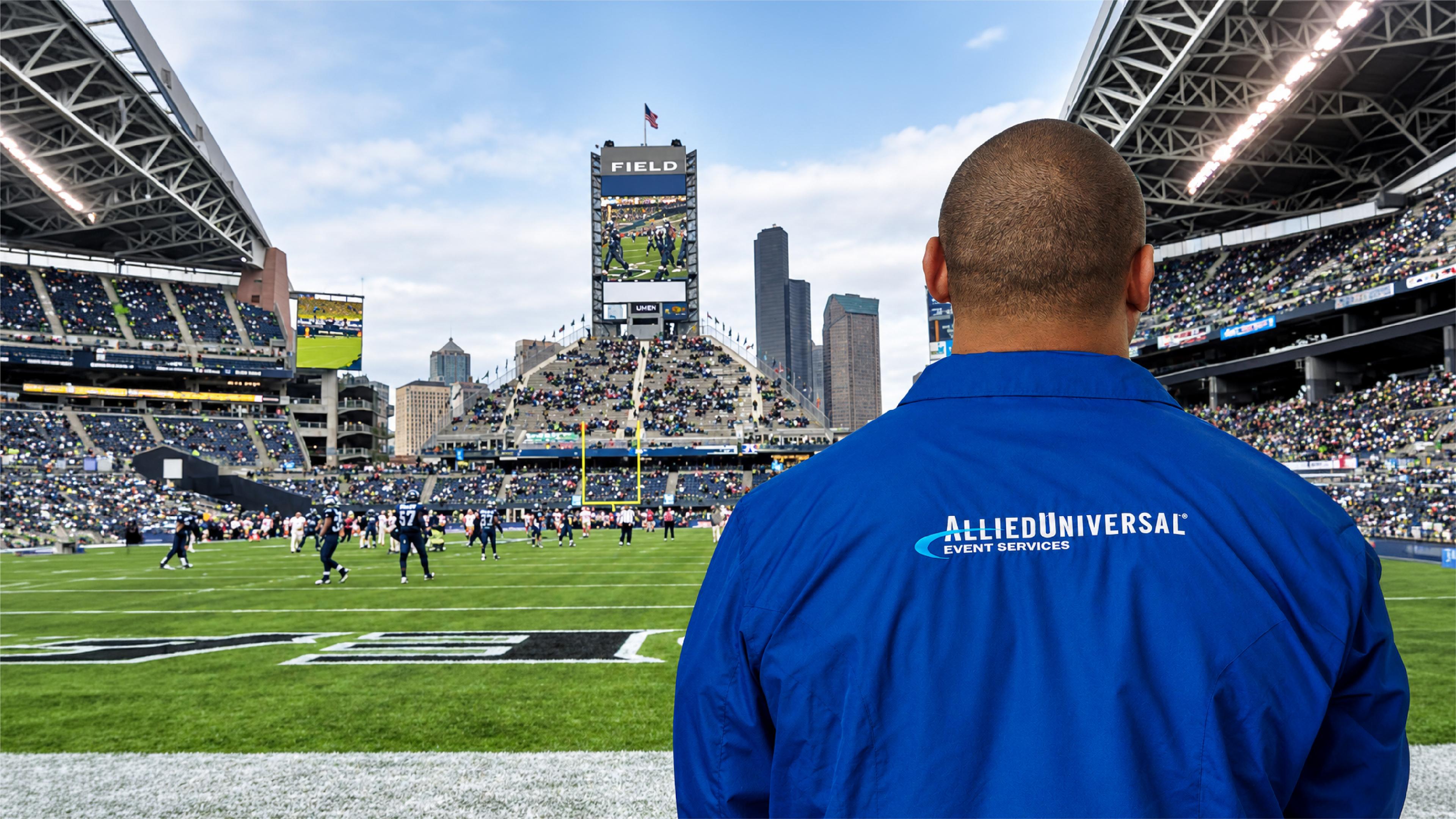 An Allied Universal Event Services employee overlooks the football field during a game at a packed stadium, with players and spectators in the background.