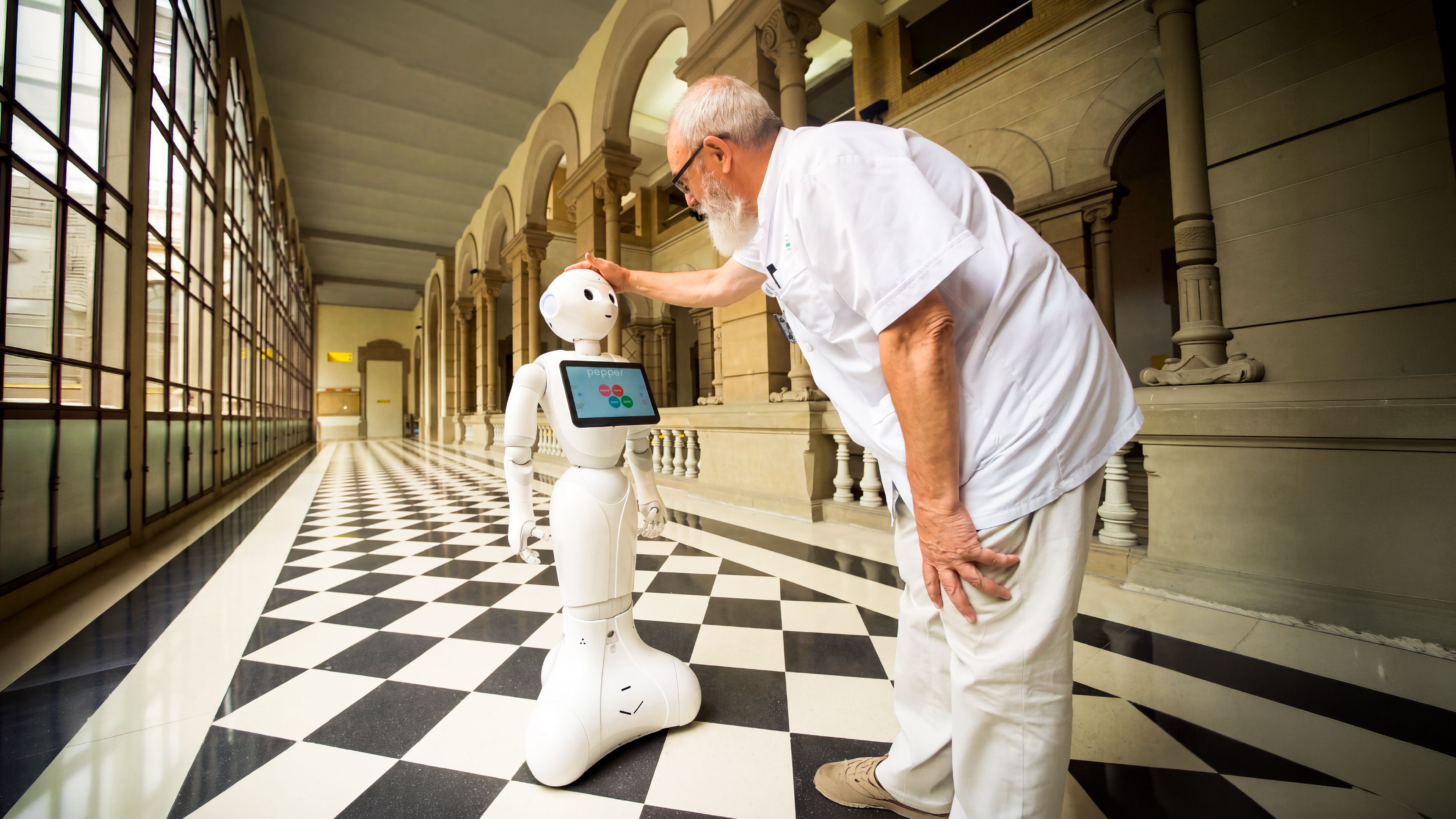 A carer greets Pepper, a humanoid service robot, in a hospital corridor