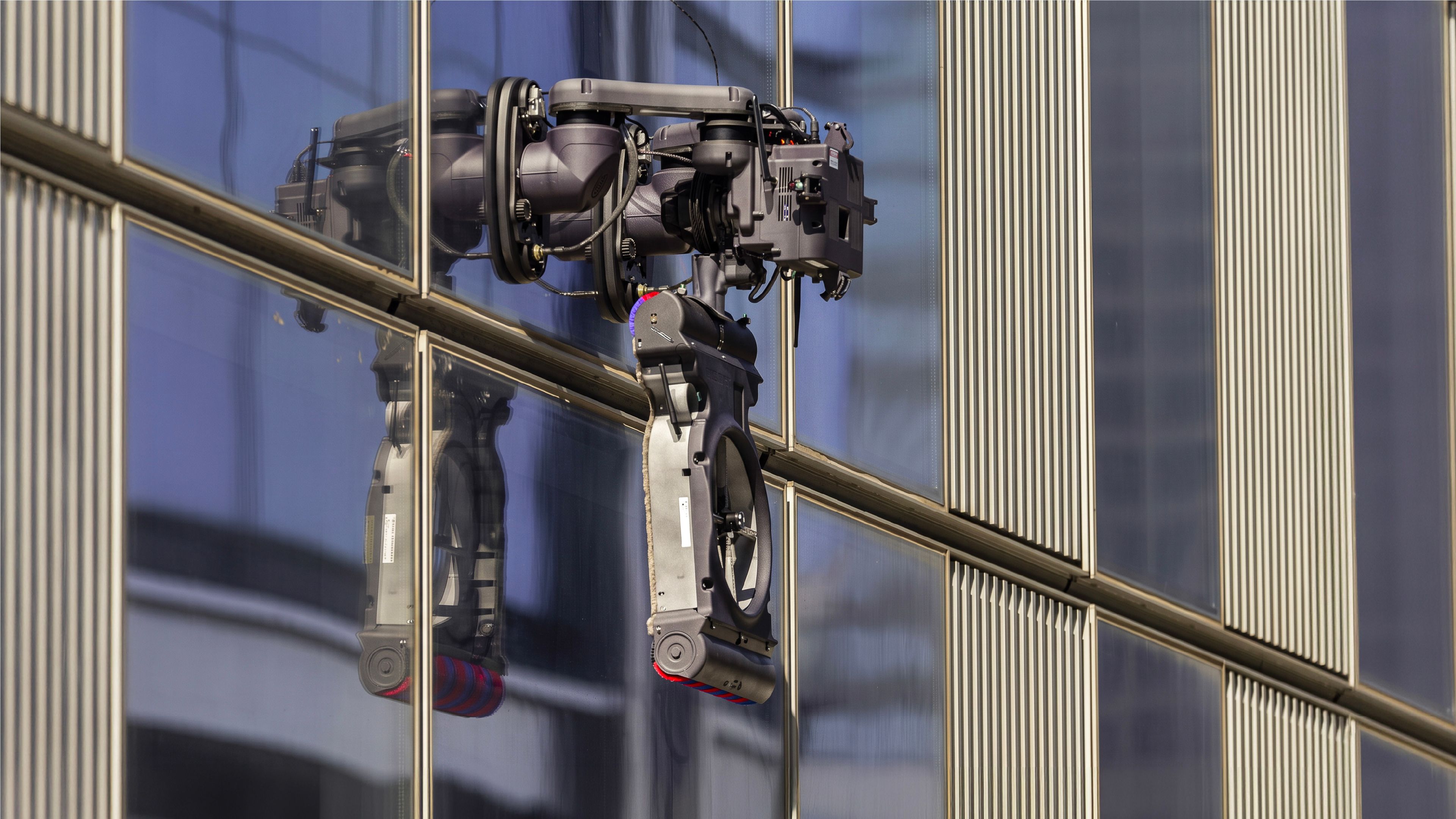 A robotic window-cleaning arm with a rotating cleaning pad and brush attachment clings to the glass facade of a modern high-rise building, its multi-jointed mechanical body reflected in the window beside it.
