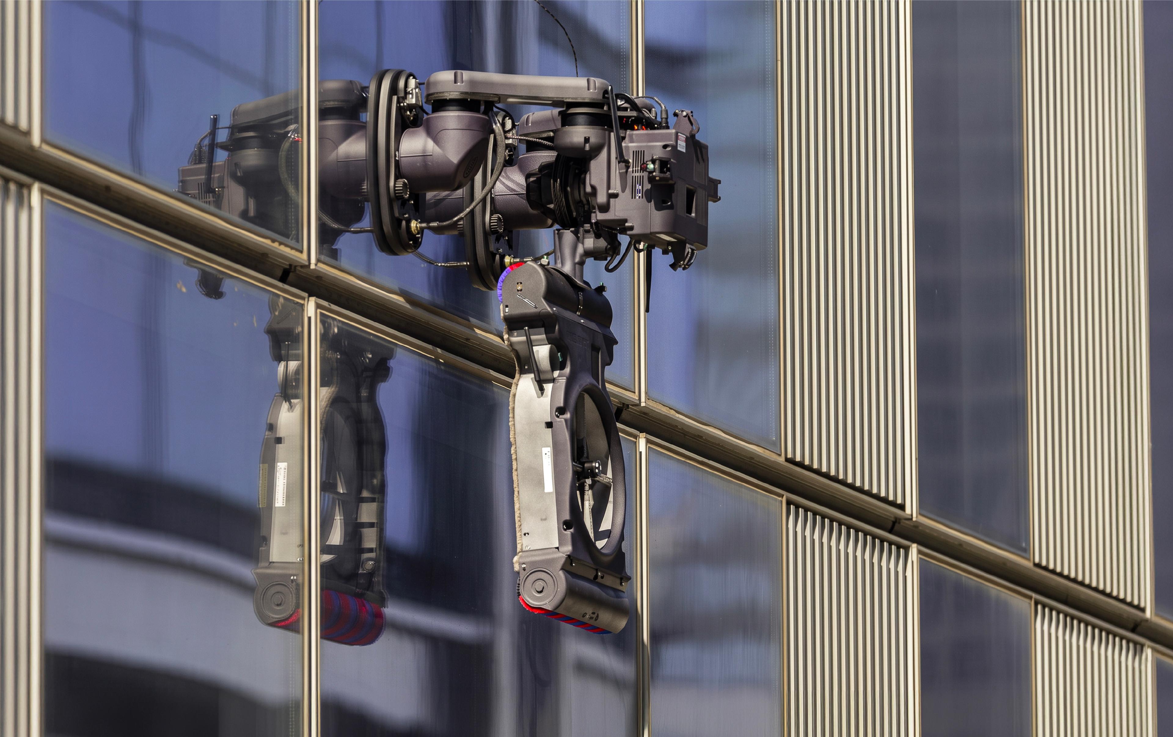 A robotic window-cleaning arm with a rotating cleaning pad and brush attachment clings to the glass facade of a modern high-rise building, its multi-jointed mechanical body reflected in the window beside it.