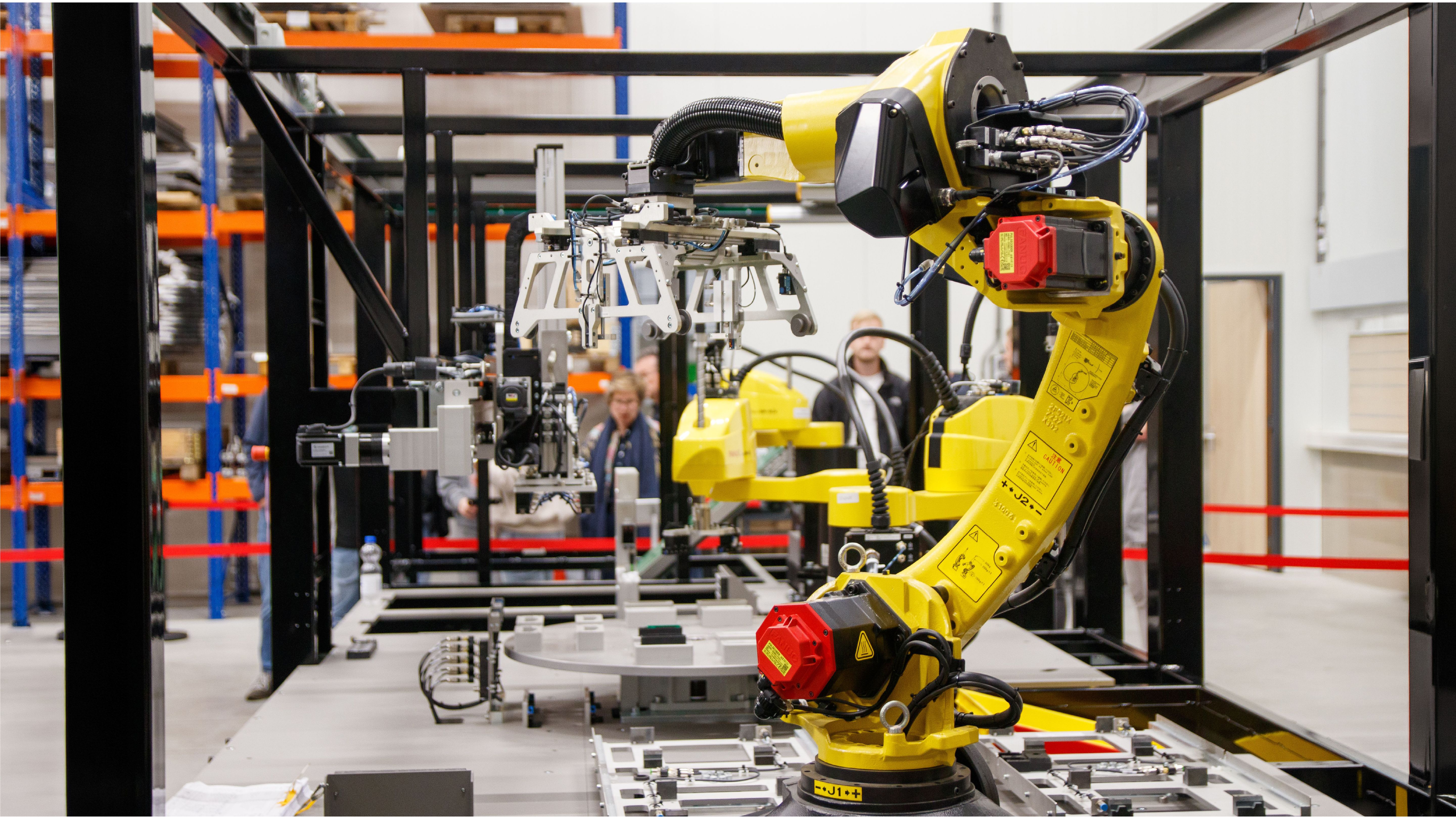 A yellow industrial robotic arm with a gripper attachment operates in a manufacturing facility, with a rotary indexing table and metal parts fixtures in the foreground and two people observing in the background, surrounded by warehouse shelving."