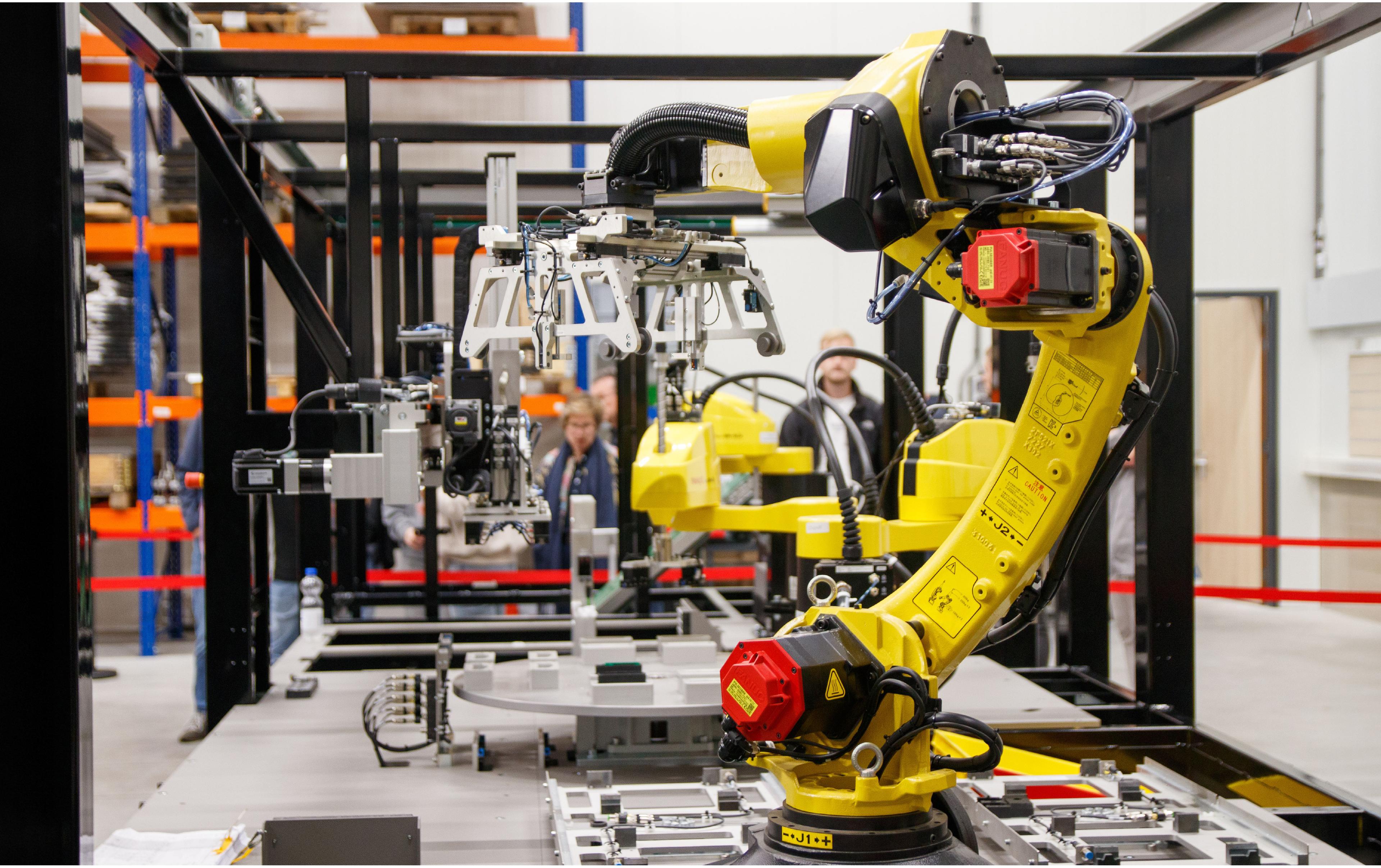 A yellow industrial robotic arm with a gripper attachment operates in a manufacturing facility, with a rotary indexing table and metal parts fixtures in the foreground and two people observing in the background, surrounded by warehouse shelving."