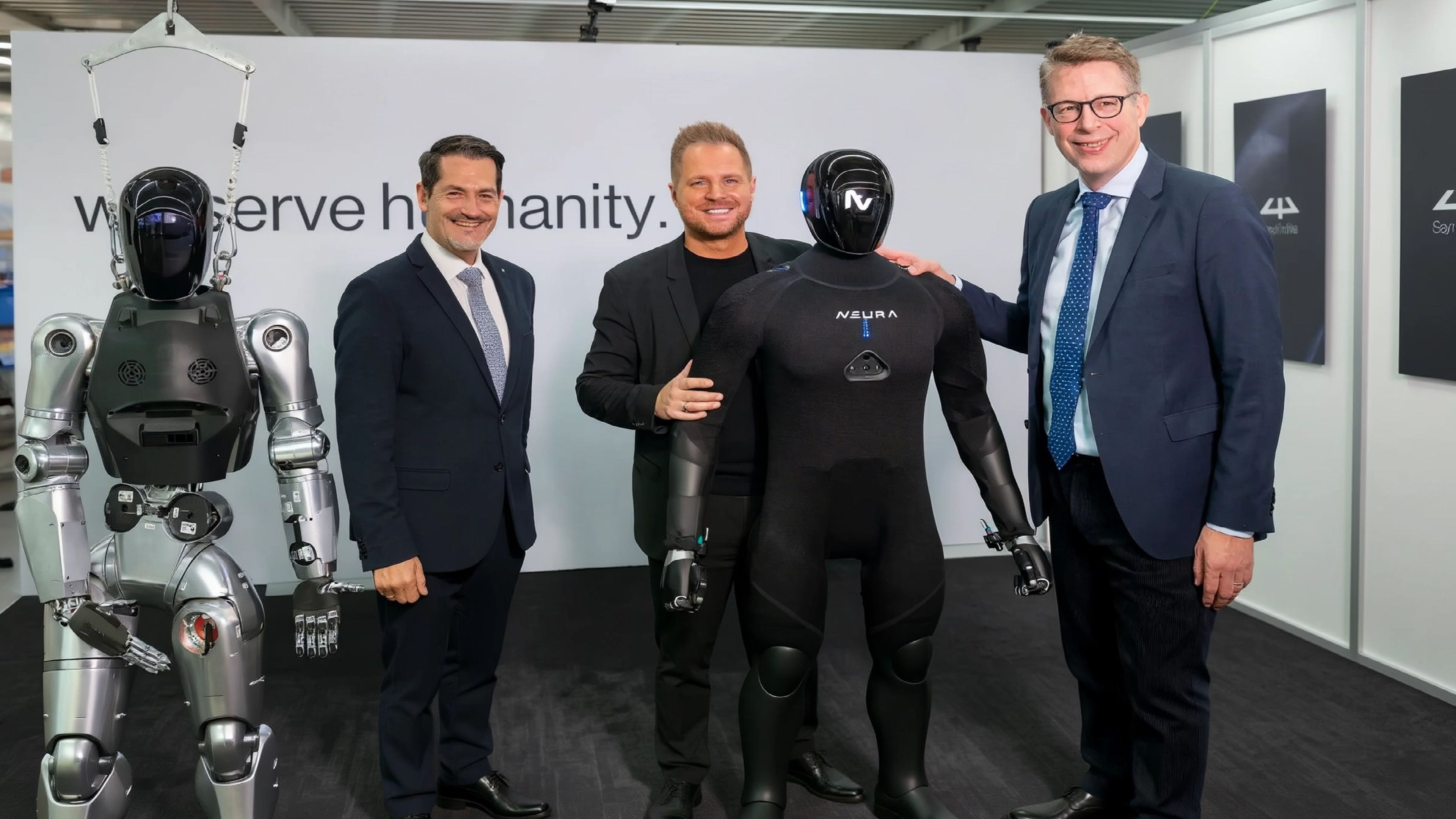 Three men pose with two generations of NEURA Robotics' humanoid robots.