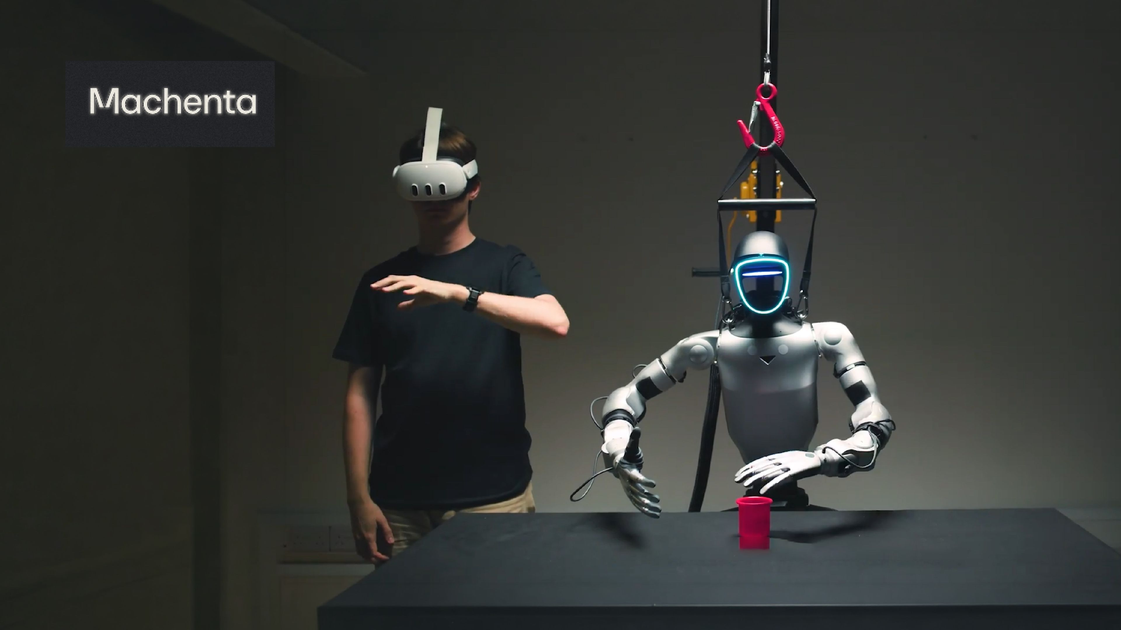 A person wearing a VR headset gestures beside a suspended humanoid robot with articulated hands during a Machenta robotics demonstration.