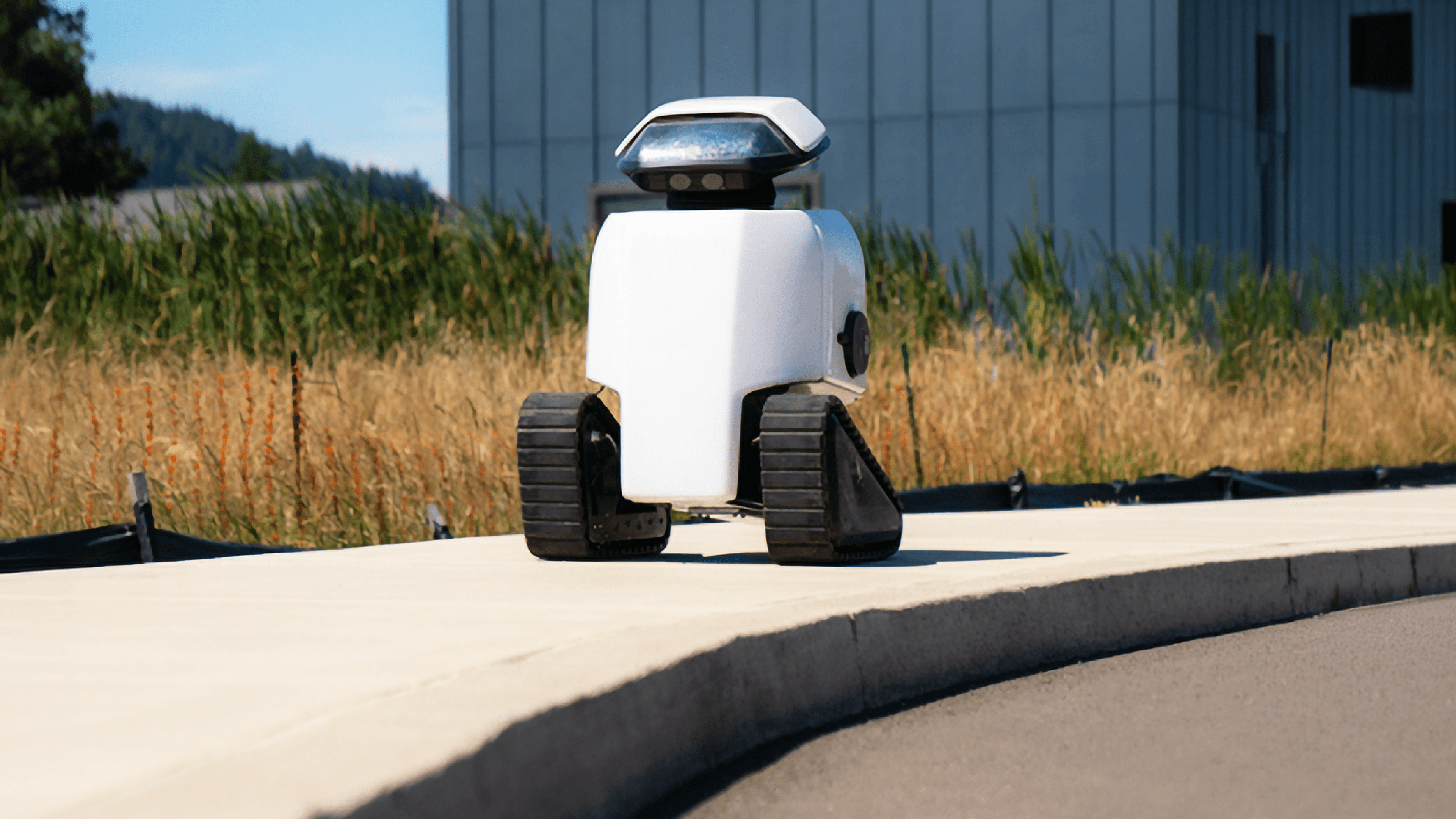 White Daxbot urban service robot with tracked wheels standing on a paved path beside dry grassland, industrial building in the background.