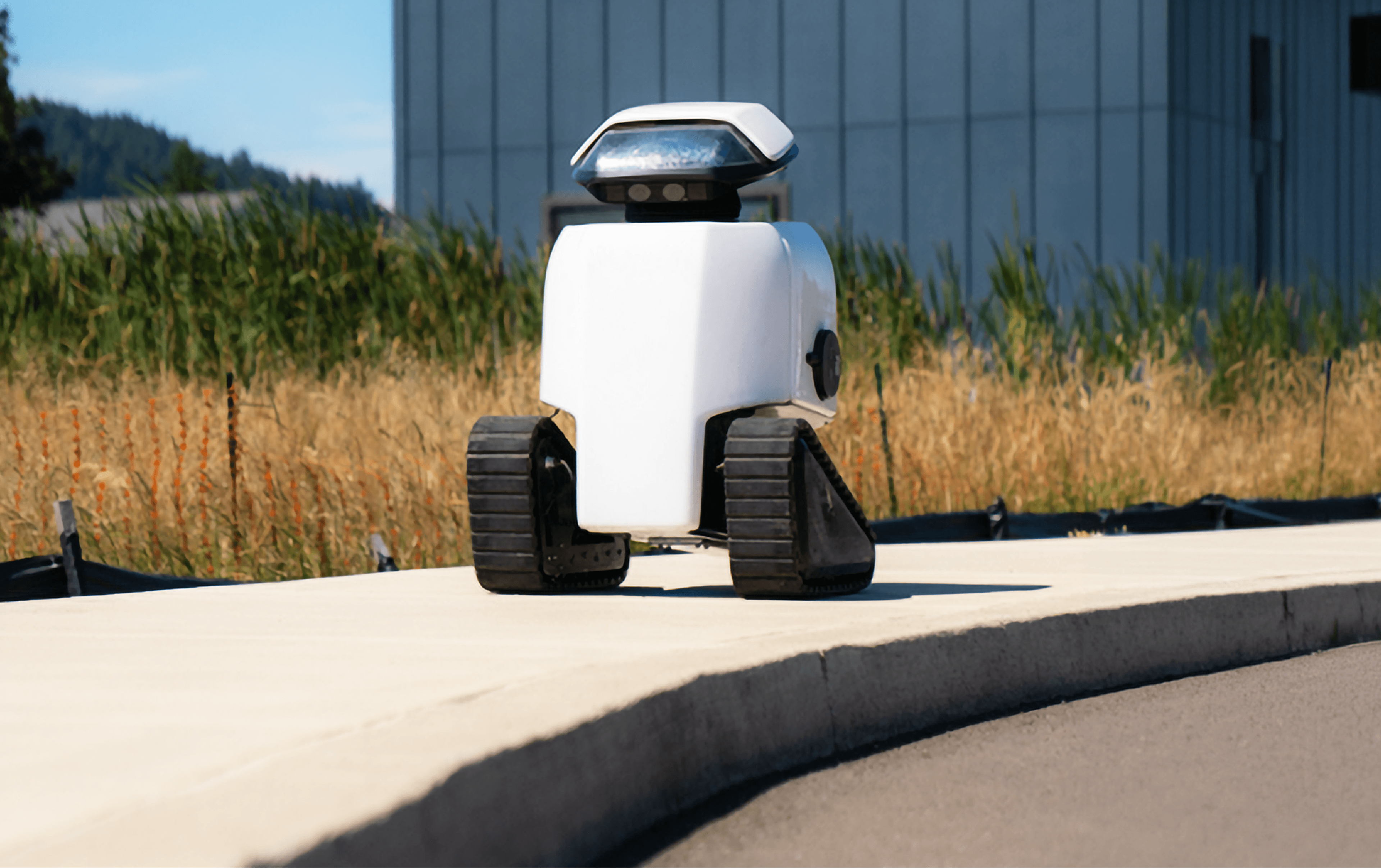 White Daxbot urban service robot with tracked wheels standing on a paved path beside dry grassland, industrial building in the background.