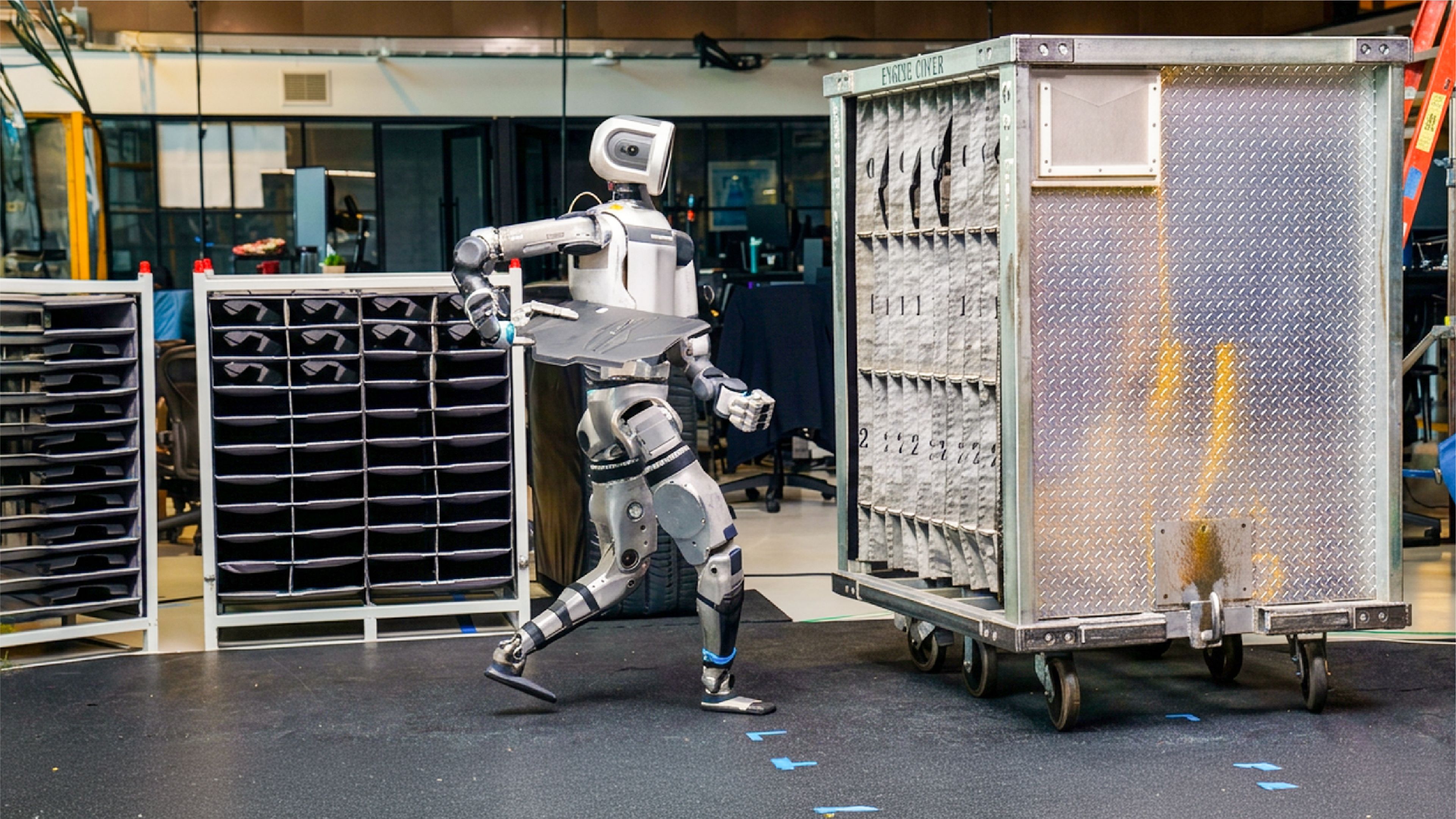 A humanoid robot carries a tray while walking between storage racks in a warehouse or workshop setting.