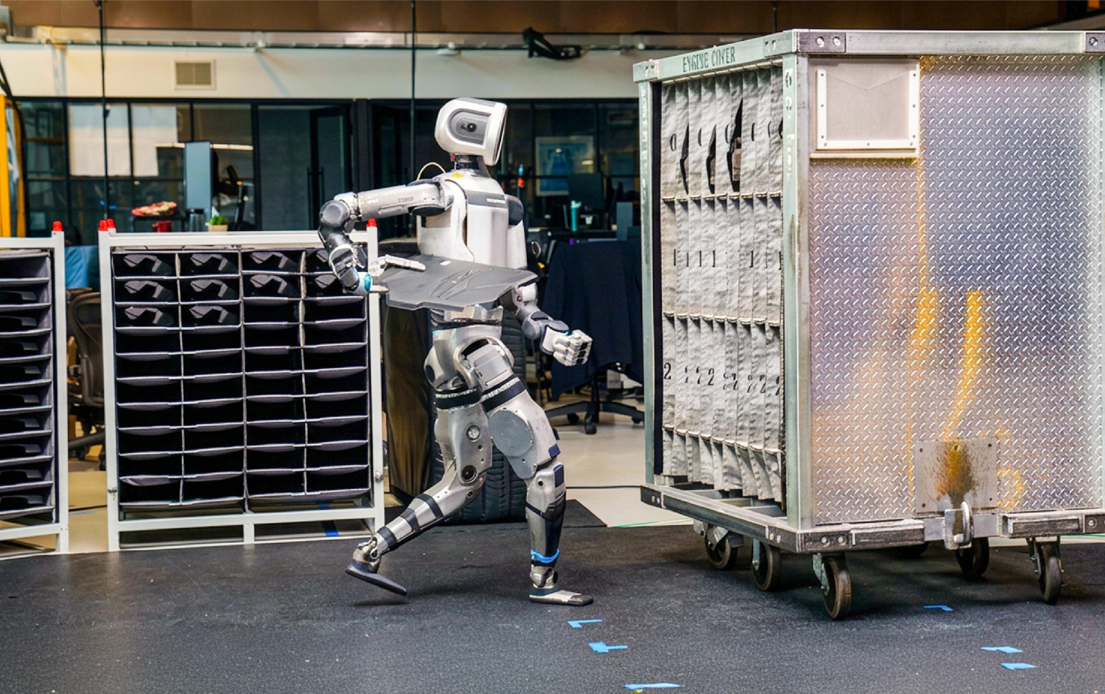 A humanoid robot carries a tray while walking between storage racks in a warehouse or workshop setting.