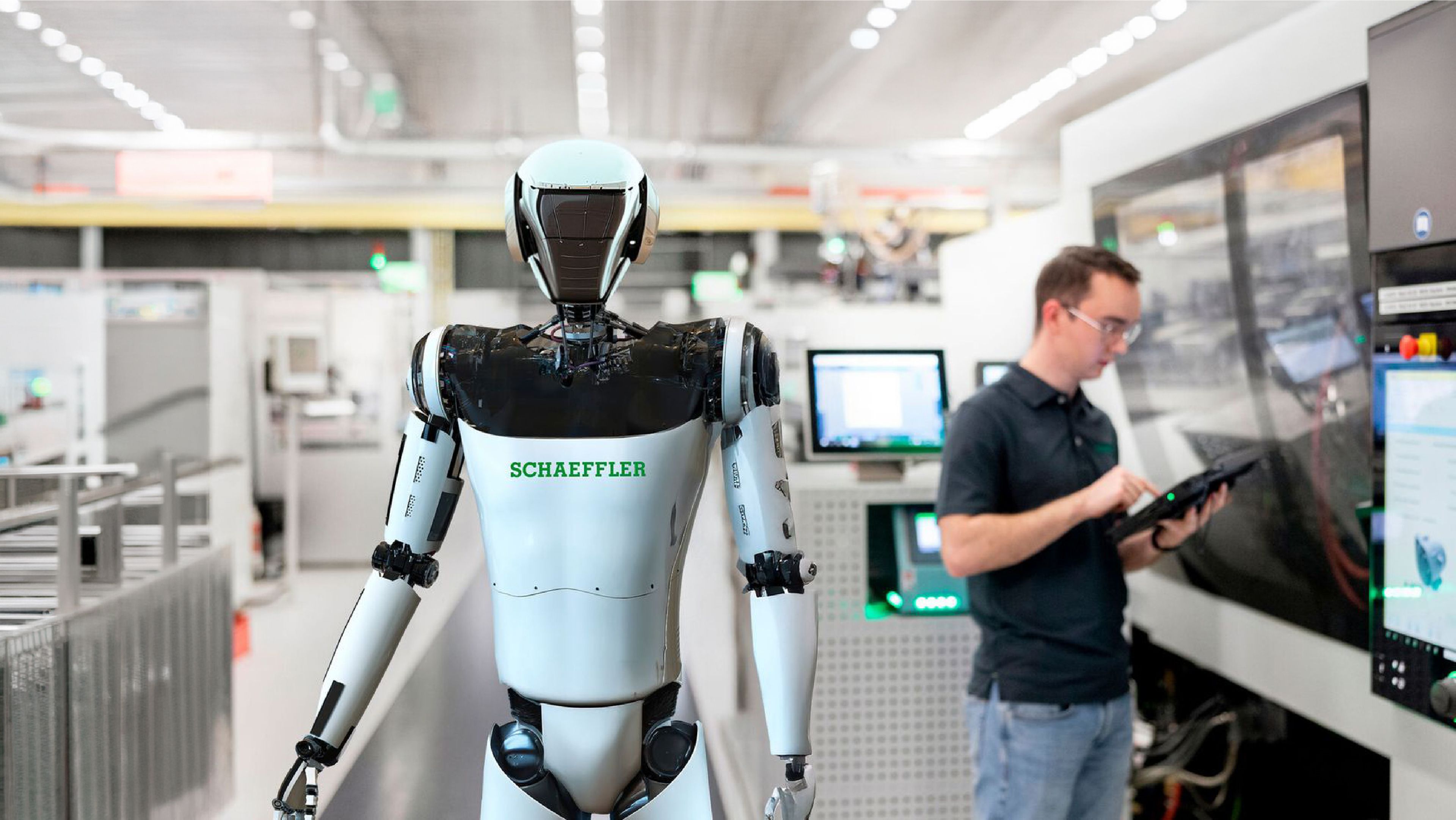 Schaeffler-branded humanoid robot standing on a factory production floor beside an employee operating a tablet near industrial machinery.