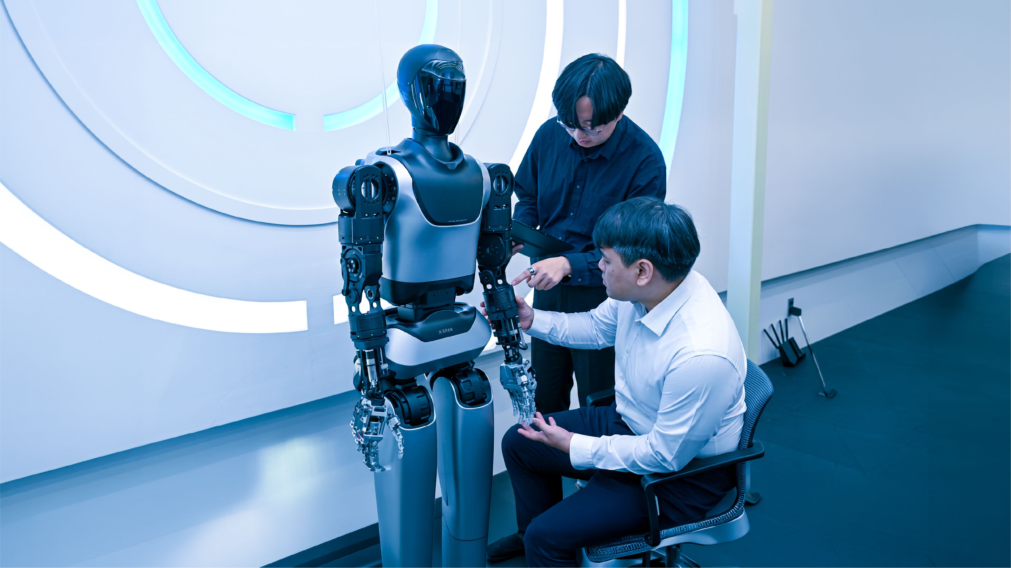 Two engineers examine the robotic hand of a humanoid robot labeled "X-SPAN" in a lab, one seated and holding its fingers, the other standing with a tablet.
