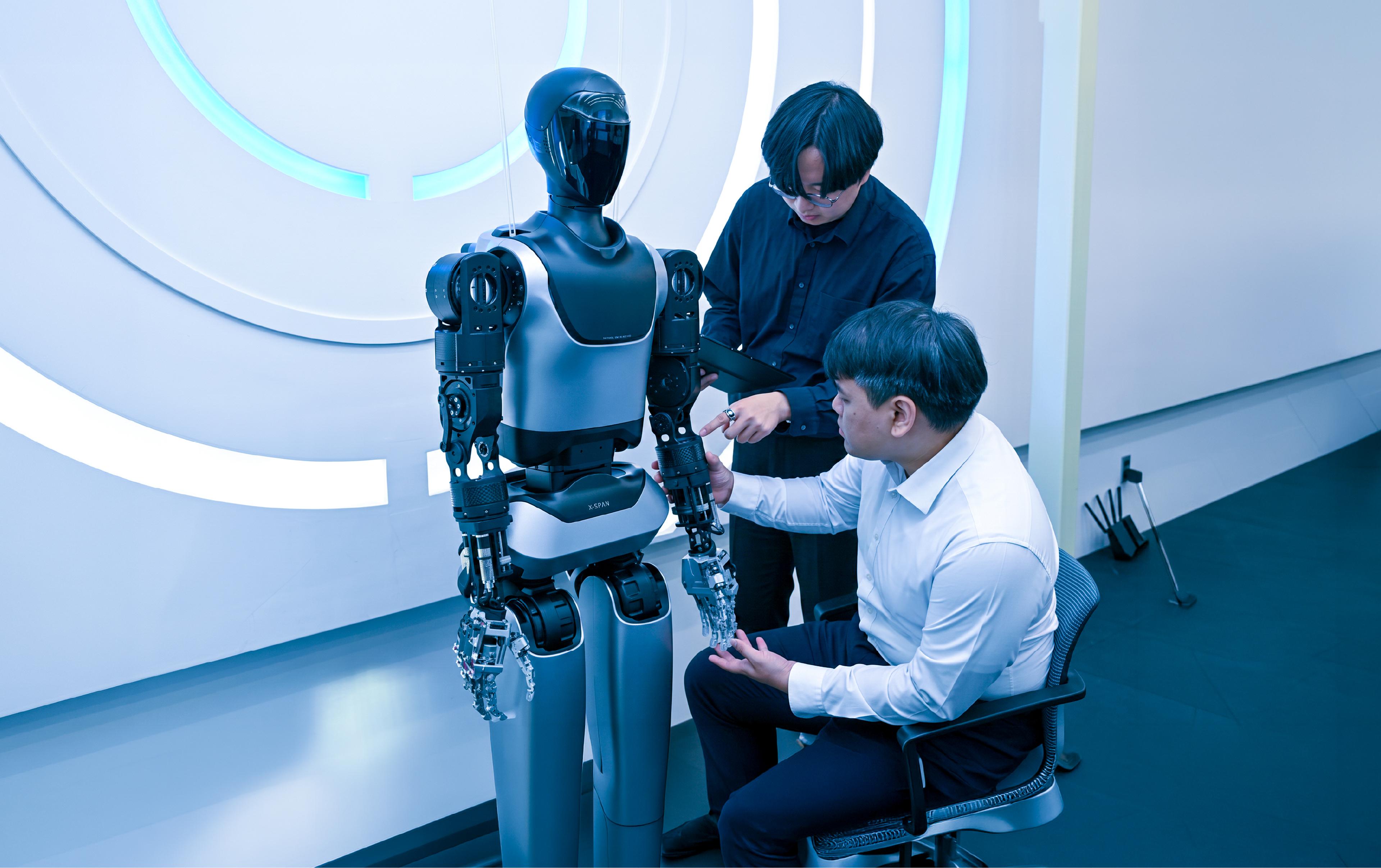 Two engineers examine the robotic hand of a humanoid robot labeled "X-SPAN" in a lab, one seated and holding its fingers, the other standing with a tablet.