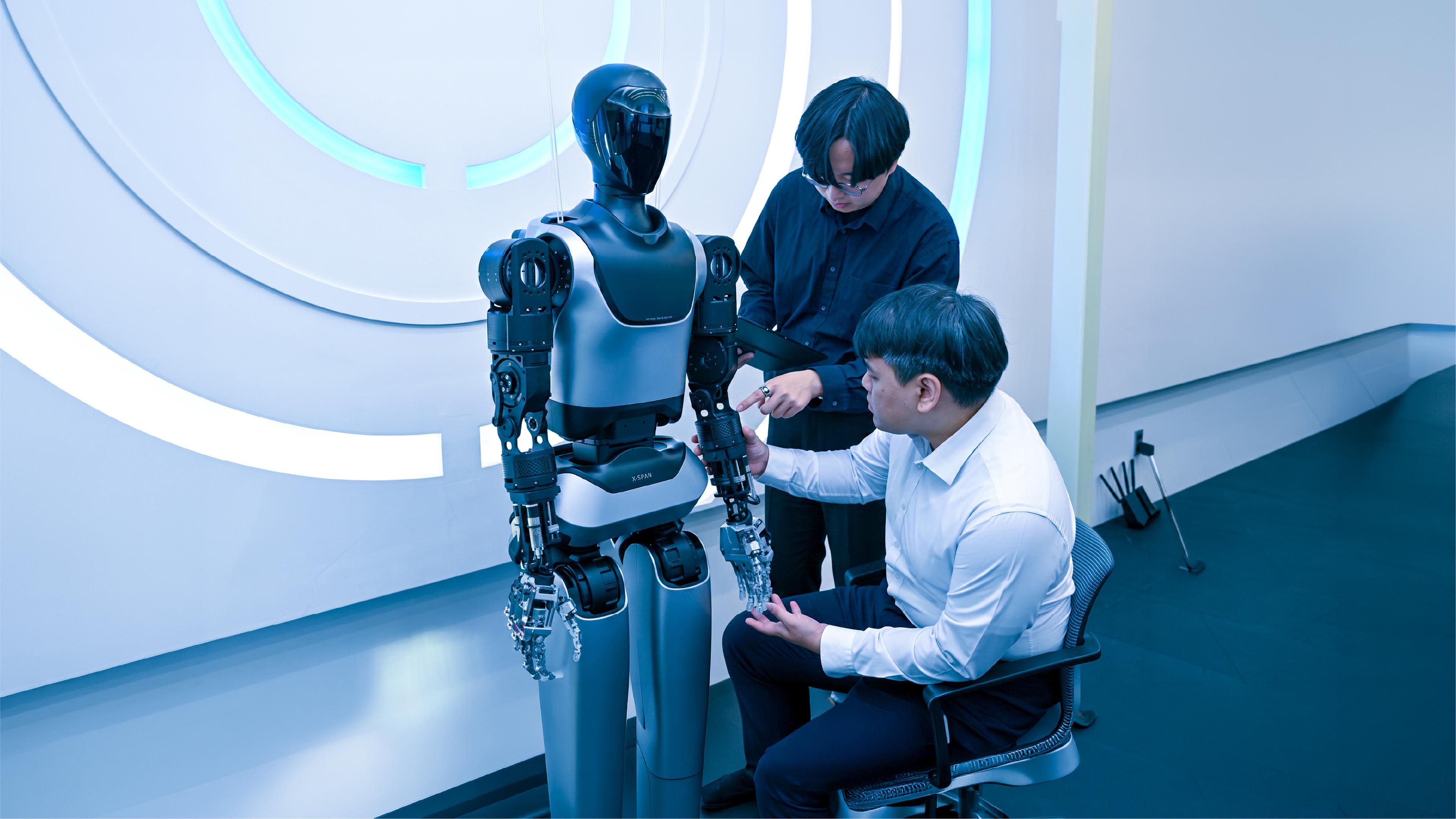 Two engineers examine the robotic hand of a humanoid robot labeled "X-SPAN" in a lab, one seated and holding its fingers, the other standing with a tablet.
