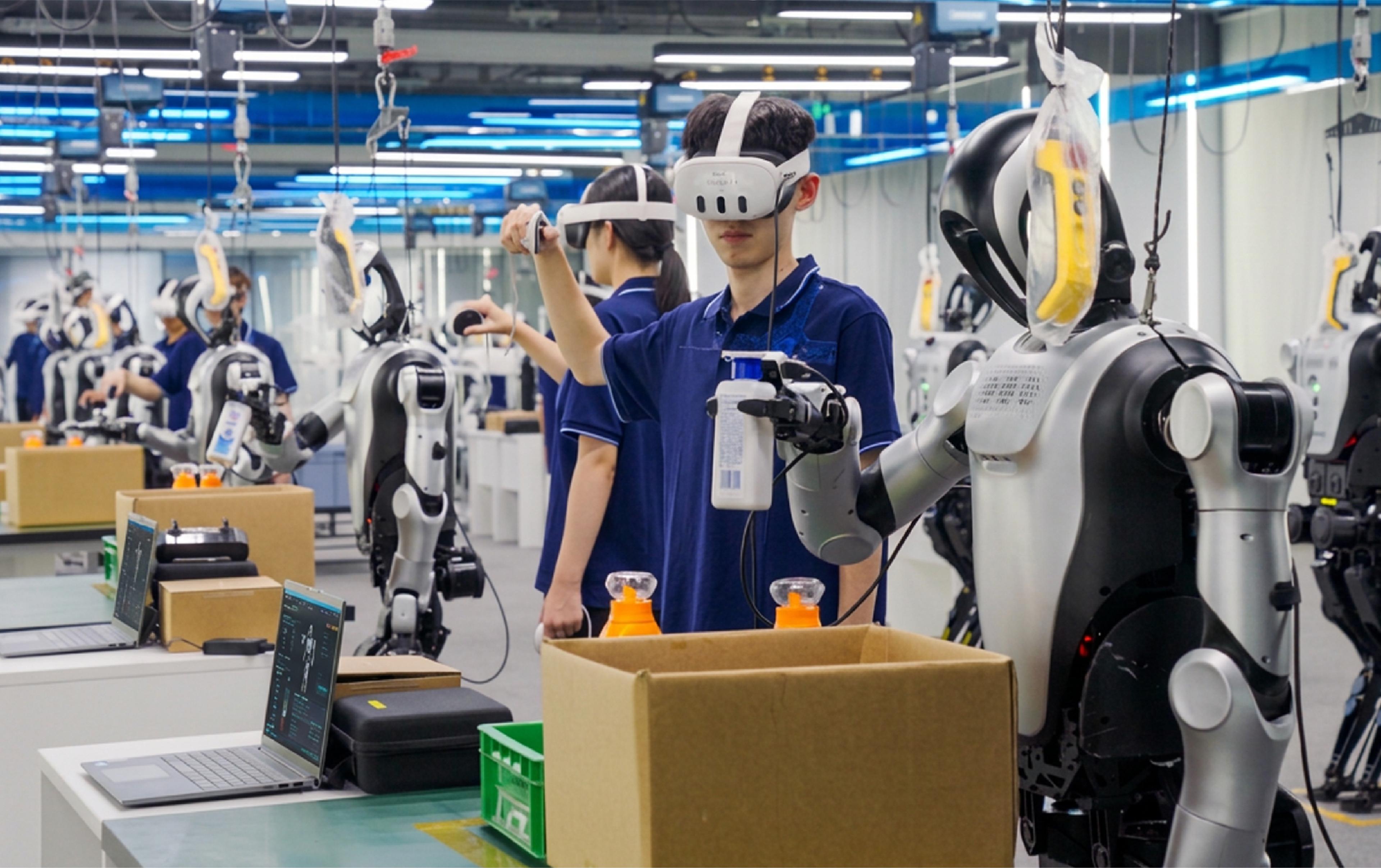 Workers wearing motion-capture headsets and controllers guide humanoid robot arms through box-packing tasks at the Shijingshan Embodied Intelligence Training Center in Beijing, with rows of suspended humanoid robots visible in the background.