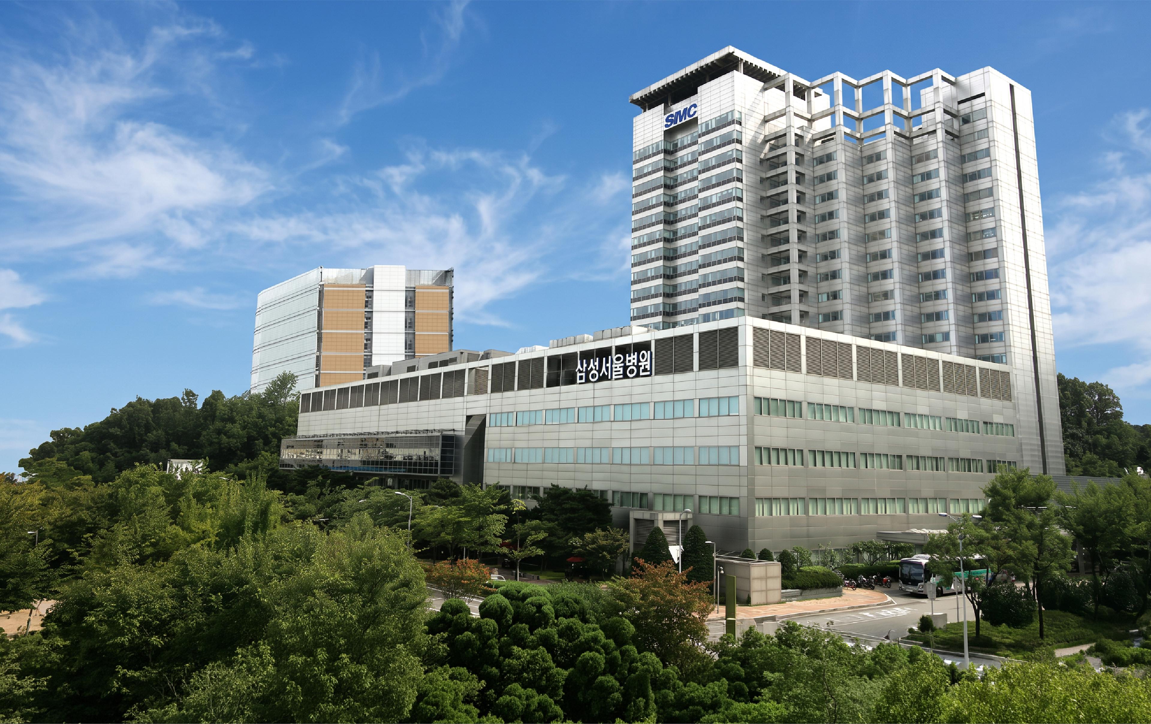 Exterior of Samsung Medical Center in Seoul, South Korea, with tall hospital buildings surrounded by trees under a blue sky.
