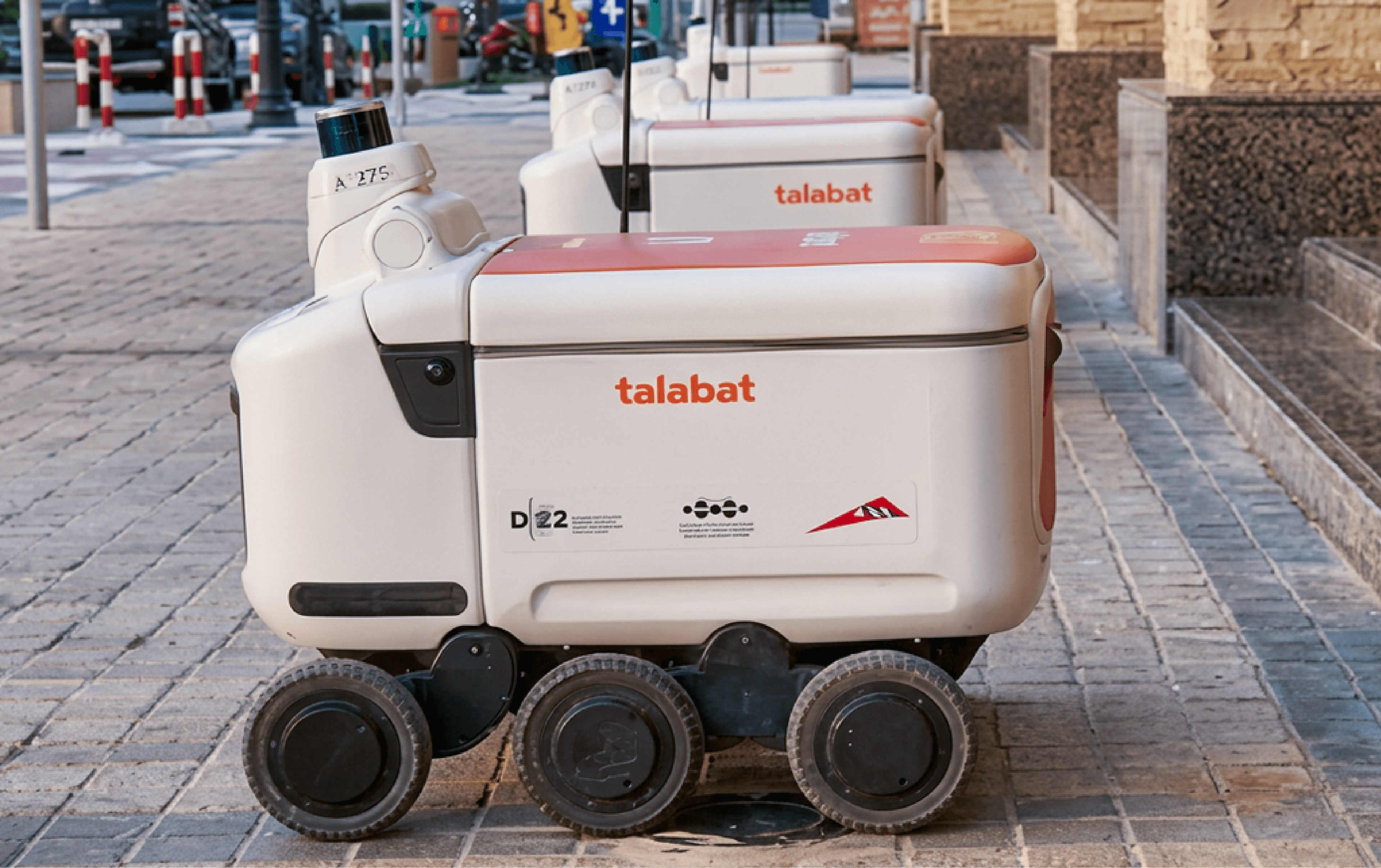 Row of six-wheeled Talabat delivery robots with orange lids parked on a paved outdoor walkway, ID marker "A275" visible on the lead unit.