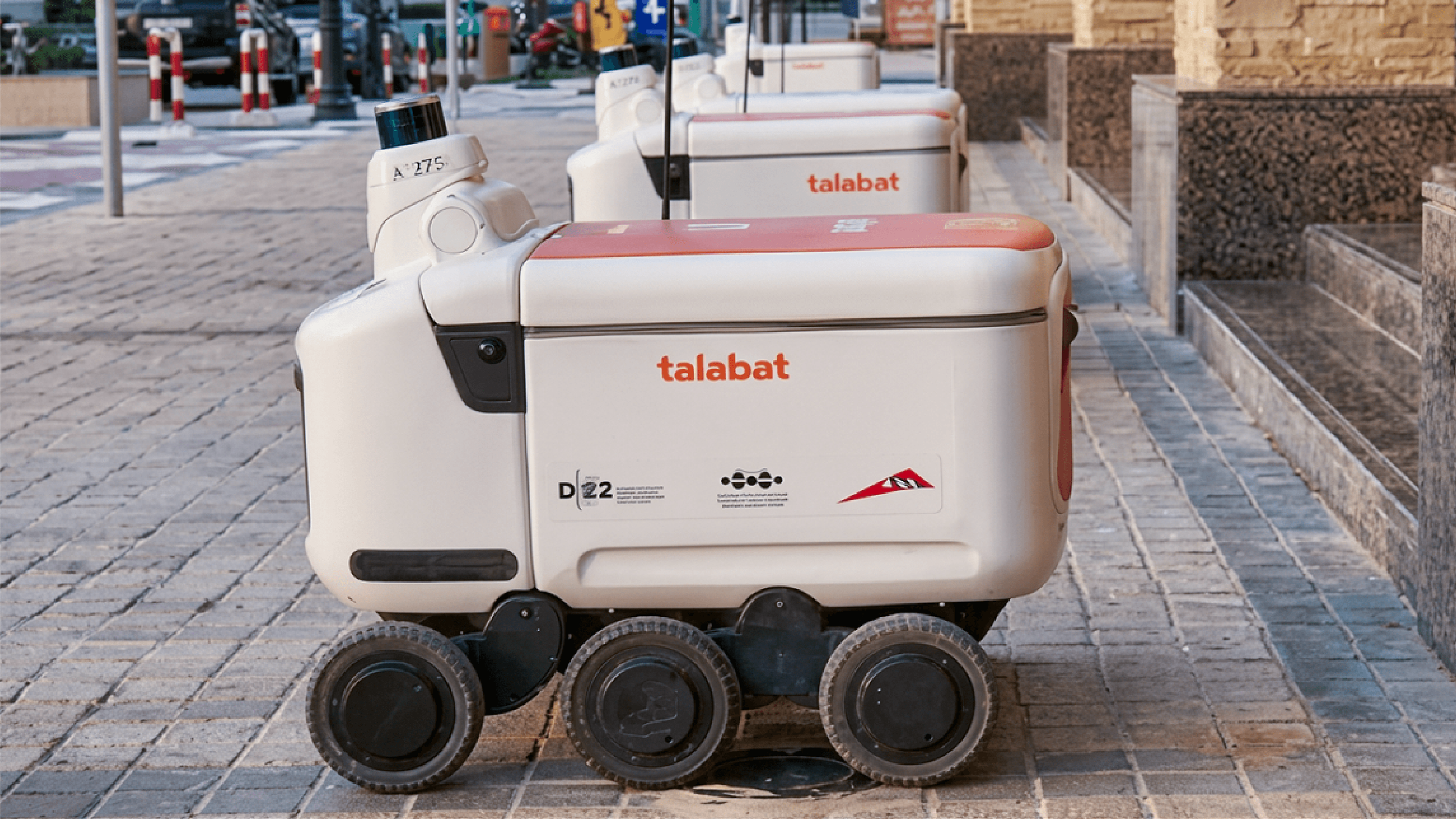Row of six-wheeled Talabat delivery robots with orange lids parked on a paved outdoor walkway, ID marker "A275" visible on the lead unit.