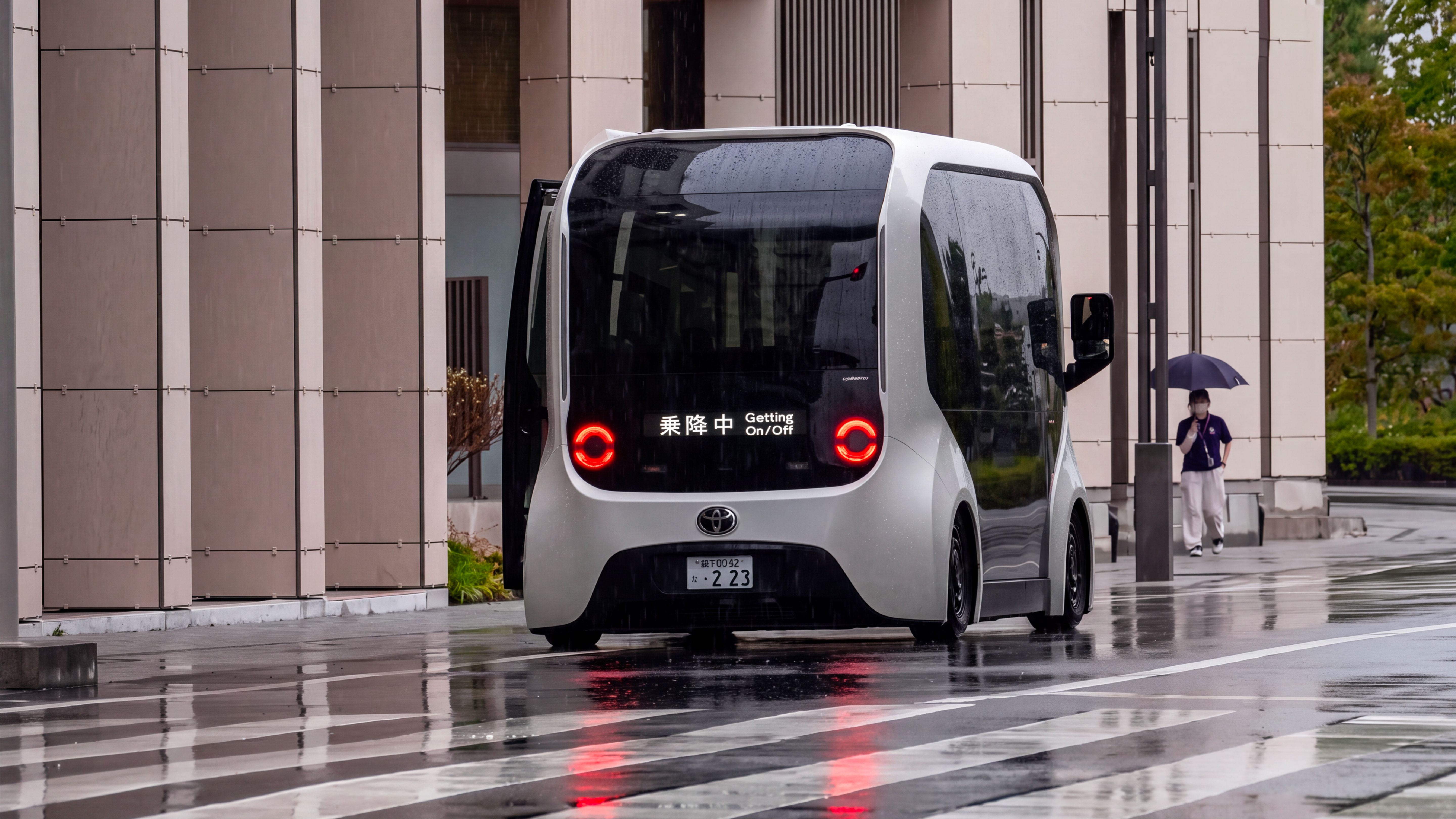 A compact autonomous shuttle stopped on a wet street outside a modern building in Japan, with a pedestrian holding an umbrella in the background.