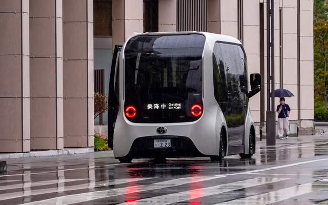 A compact autonomous shuttle stopped on a wet street outside a modern building in Japan, with a pedestrian holding an umbrella in the background.
