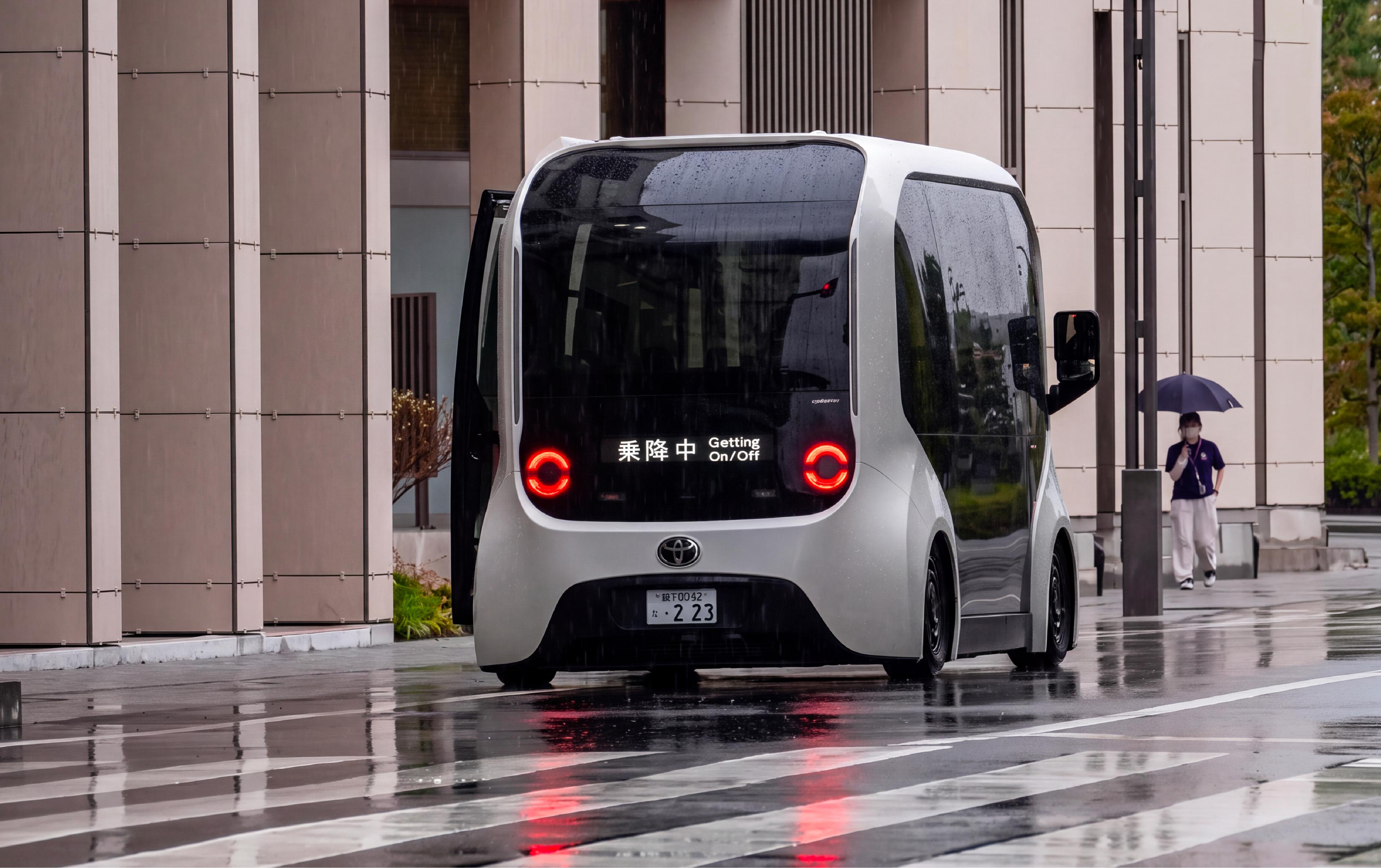 A compact autonomous shuttle stopped on a wet street outside a modern building in Japan, with a pedestrian holding an umbrella in the background.