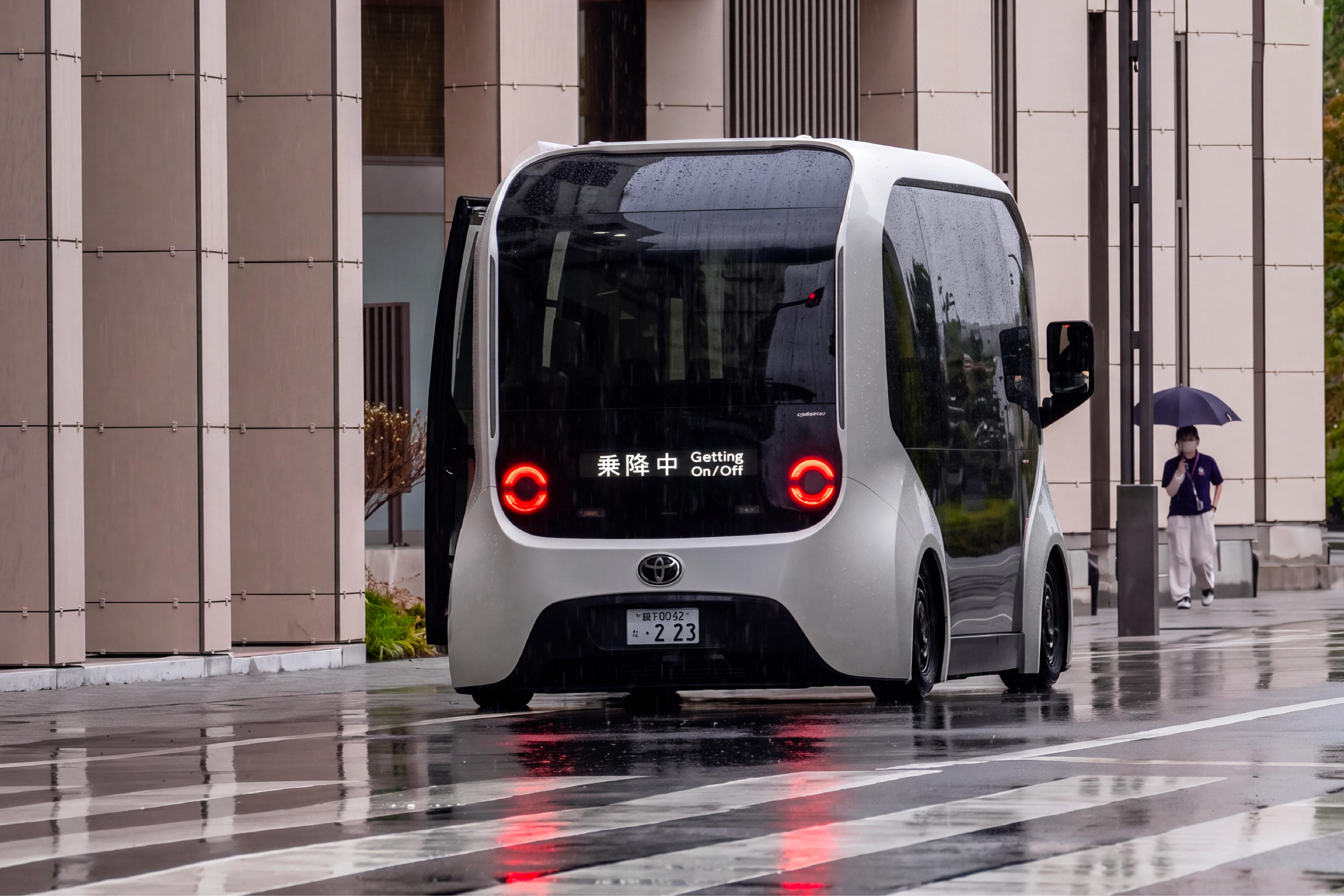 A compact autonomous shuttle stopped on a wet street outside a modern building in Japan, with a pedestrian holding an umbrella in the background.
