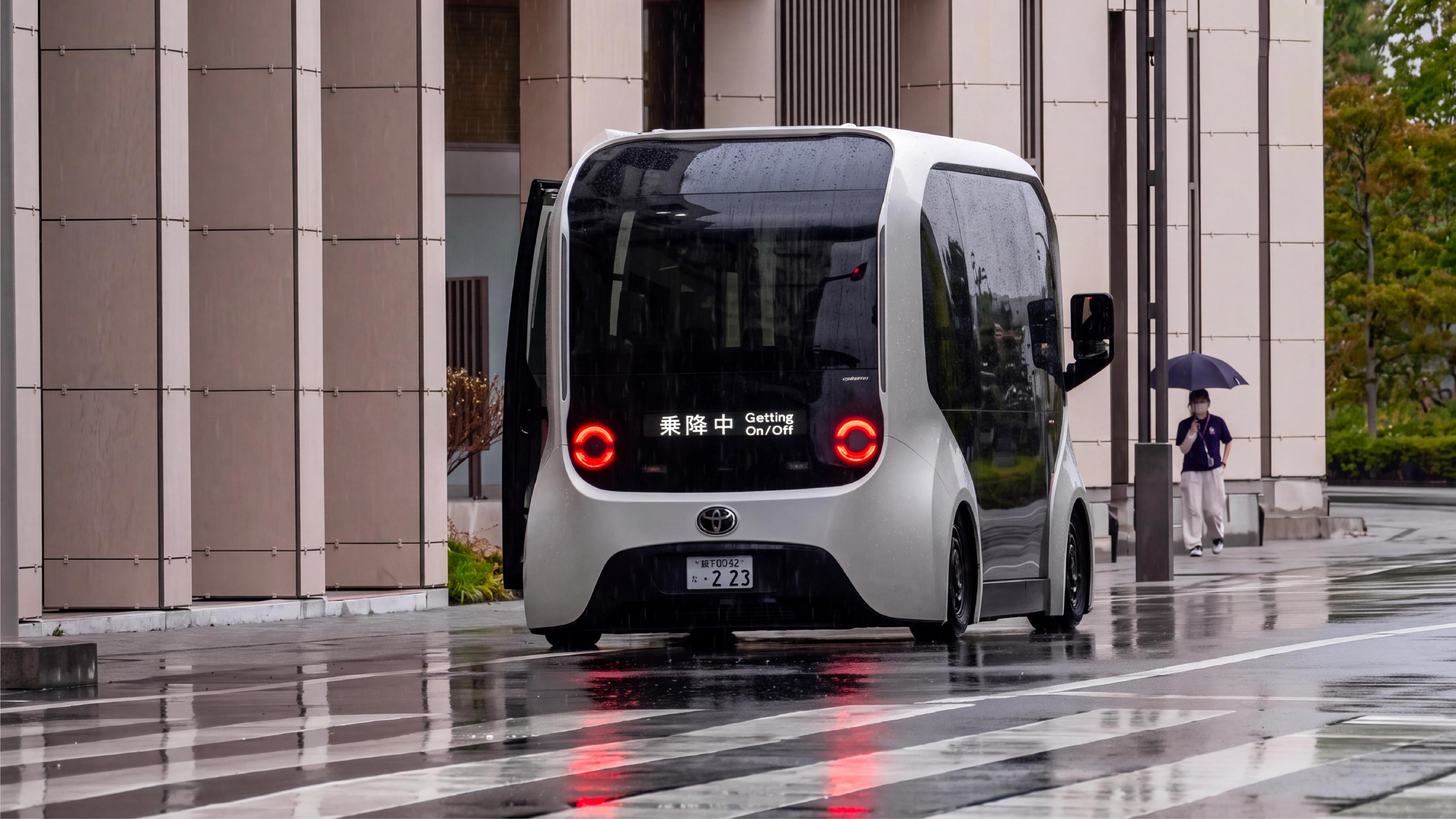 A compact autonomous shuttle stopped on a wet street outside a modern building in Japan, with a pedestrian holding an umbrella in the background.