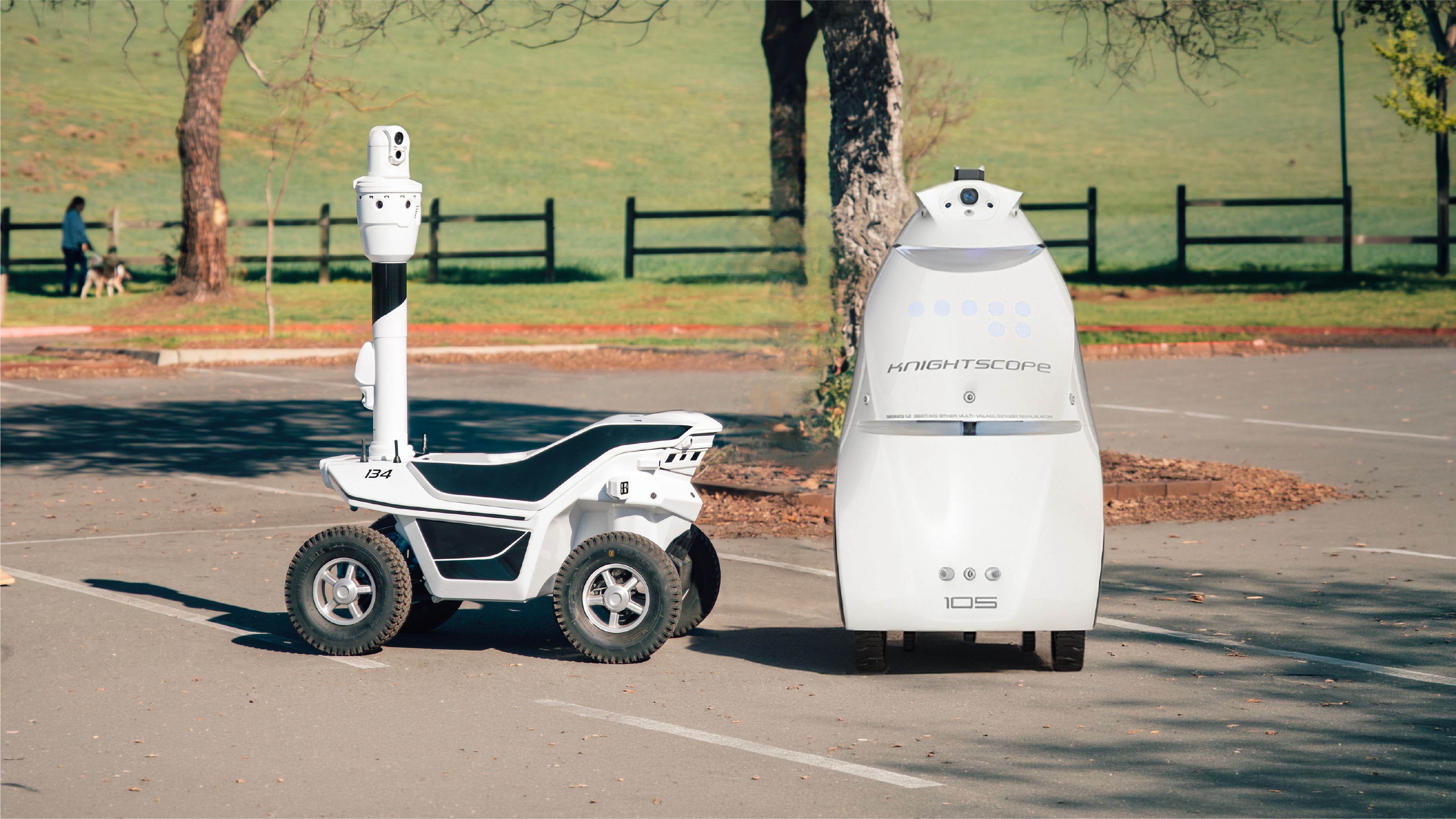 Two outdoor security patrol robots at a warehouse loading area: a compact two-wheel Ascento Guard robot in the background, and a four-wheel Artisty-branded patrol robot with a raised camera mast in the foreground."