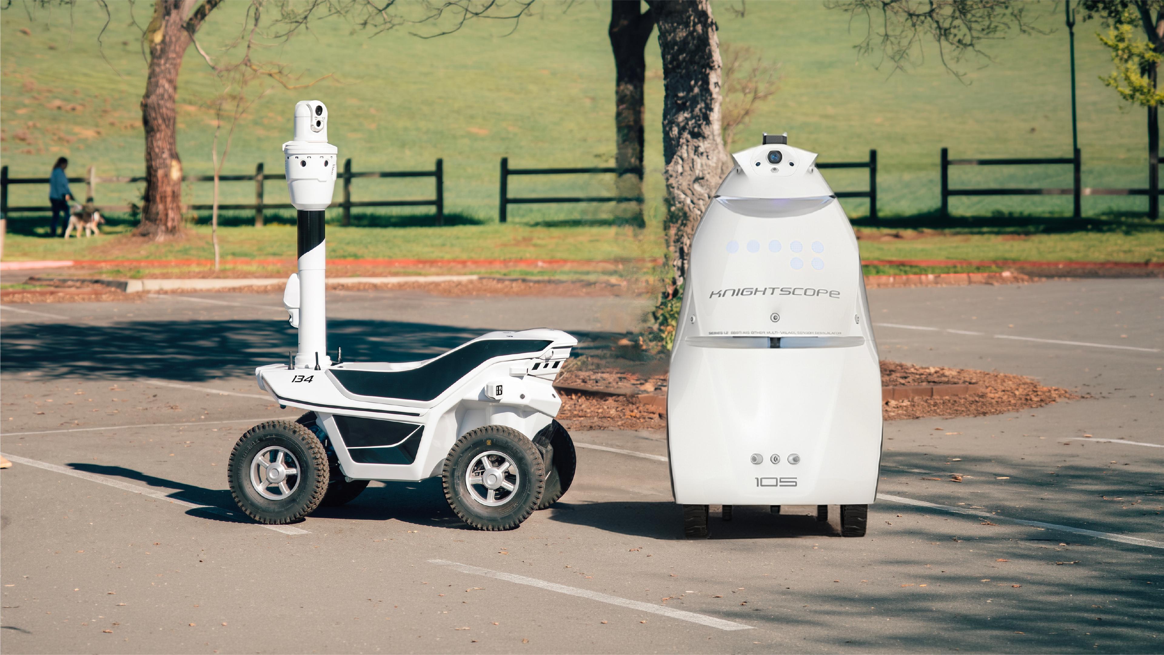 Two outdoor security patrol robots at a warehouse loading area: a compact two-wheel Ascento Guard robot in the background, and a four-wheel Artisty-branded patrol robot with a raised camera mast in the foreground."
