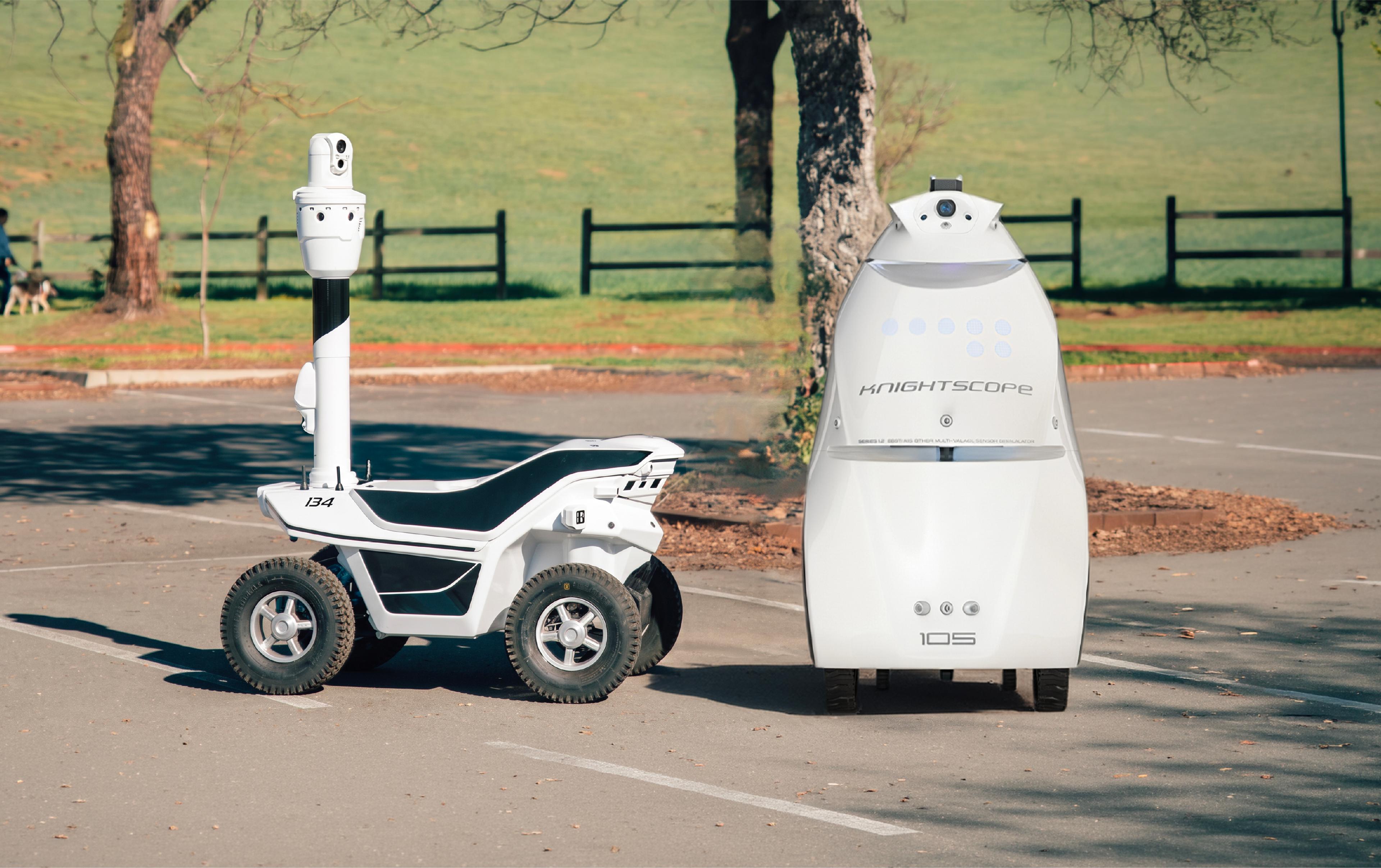 Two outdoor security patrol robots at a warehouse loading area: a compact two-wheel Ascento Guard robot in the background, and a four-wheel Artisty-branded patrol robot with a raised camera mast in the foreground."