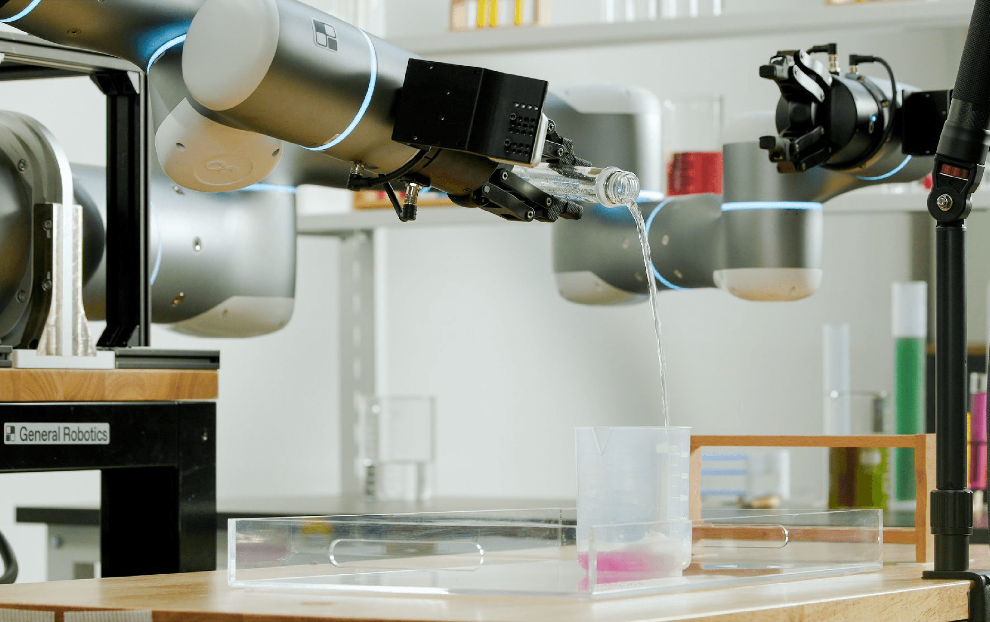 Two robot arms demonstrate laboratory liquid handling as one pours water from a glass bottle into a measuring cup.