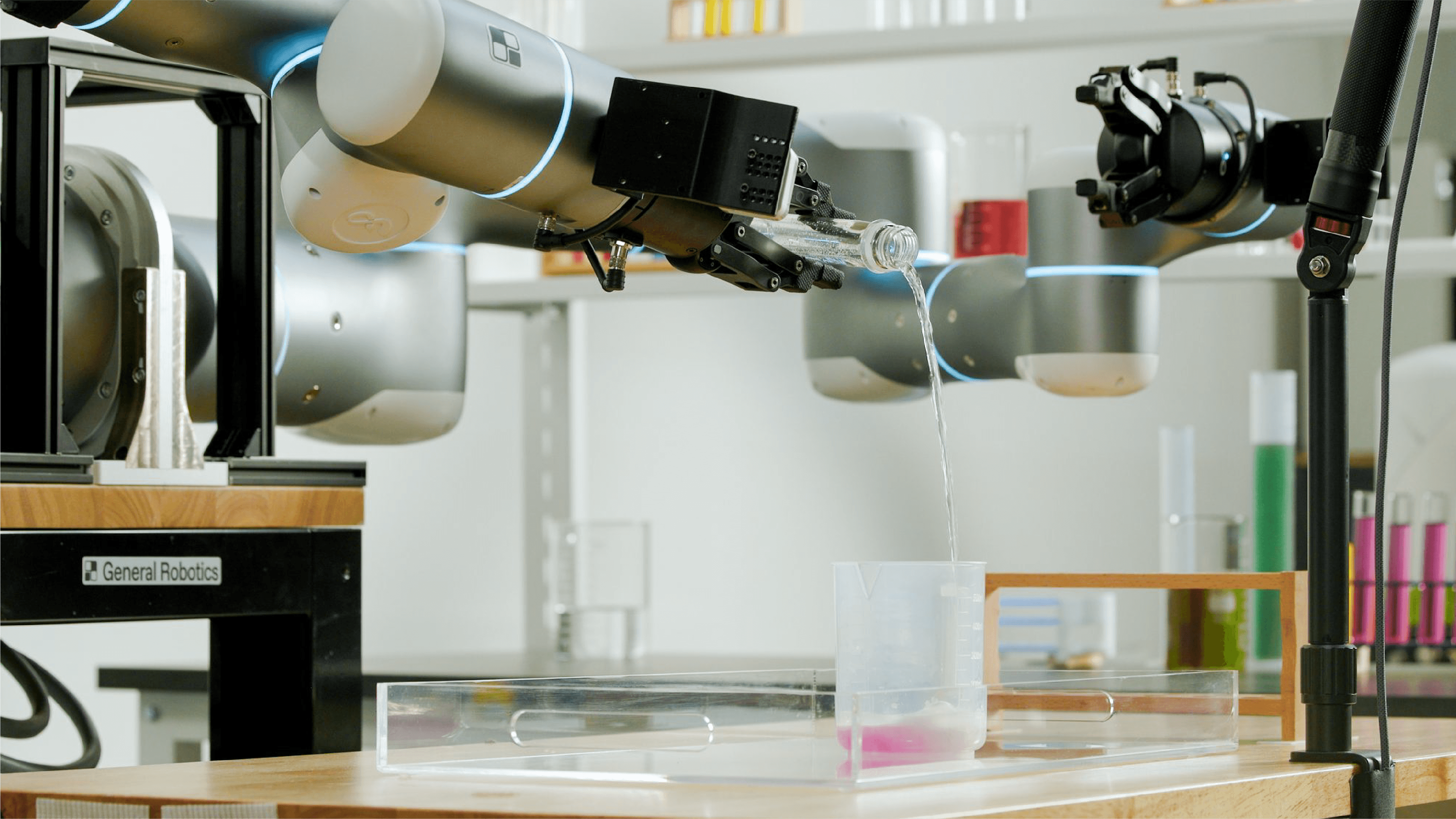 Two robot arms demonstrate laboratory liquid handling as one pours water from a glass bottle into a measuring cup.