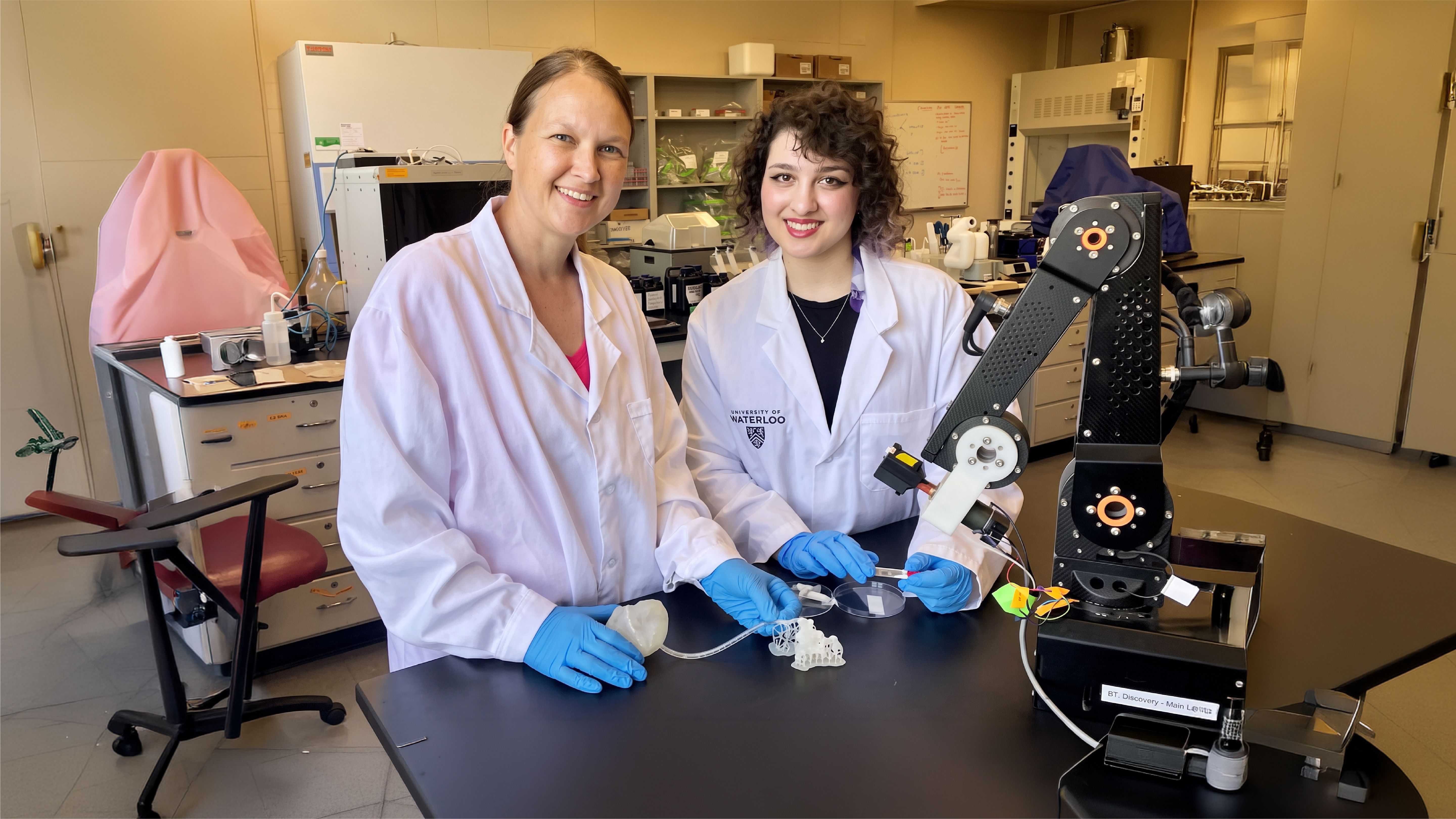 Two women in white lab coats and blue gloves stand at a lab bench beside a black robotic arm, one holding a petri dish and a 3D-printed lattice sample.
