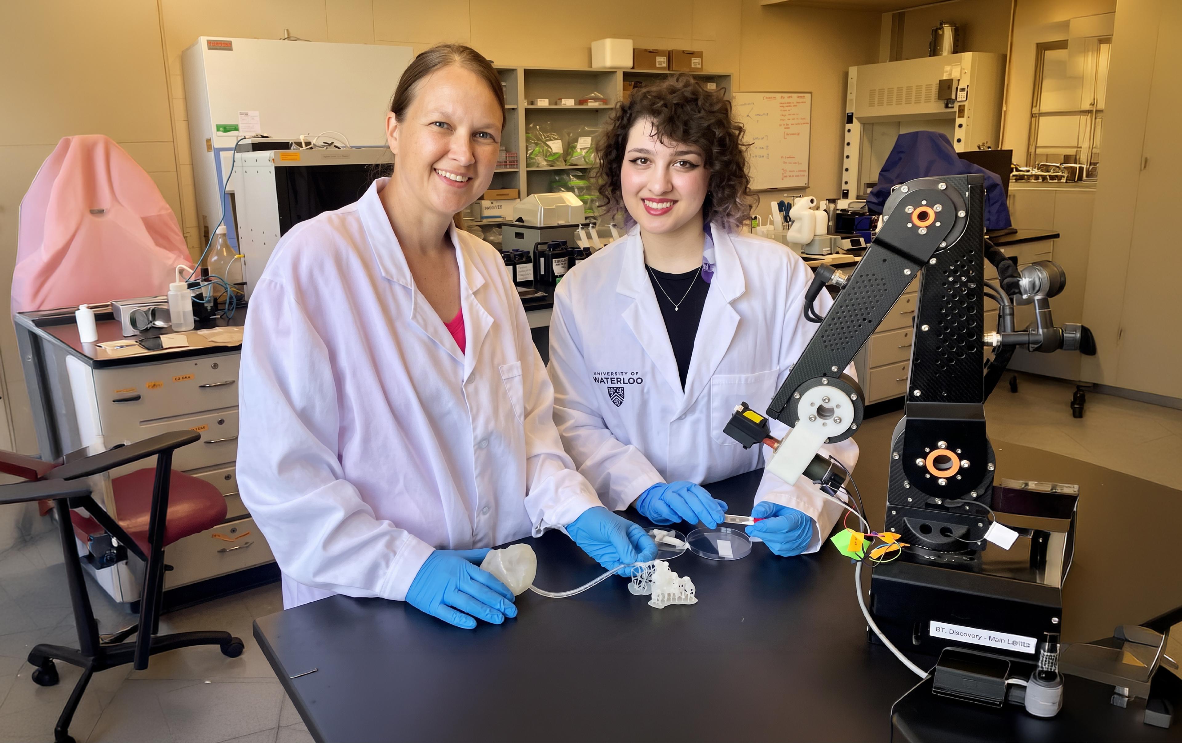 Two women in white lab coats and blue gloves stand at a lab bench beside a black robotic arm, one holding a petri dish and a 3D-printed lattice sample.