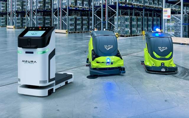 A NEURA logistics robot alongside two green and grey ADLATUS cleaning robots on a warehouse floor lined with tall storage racks.
