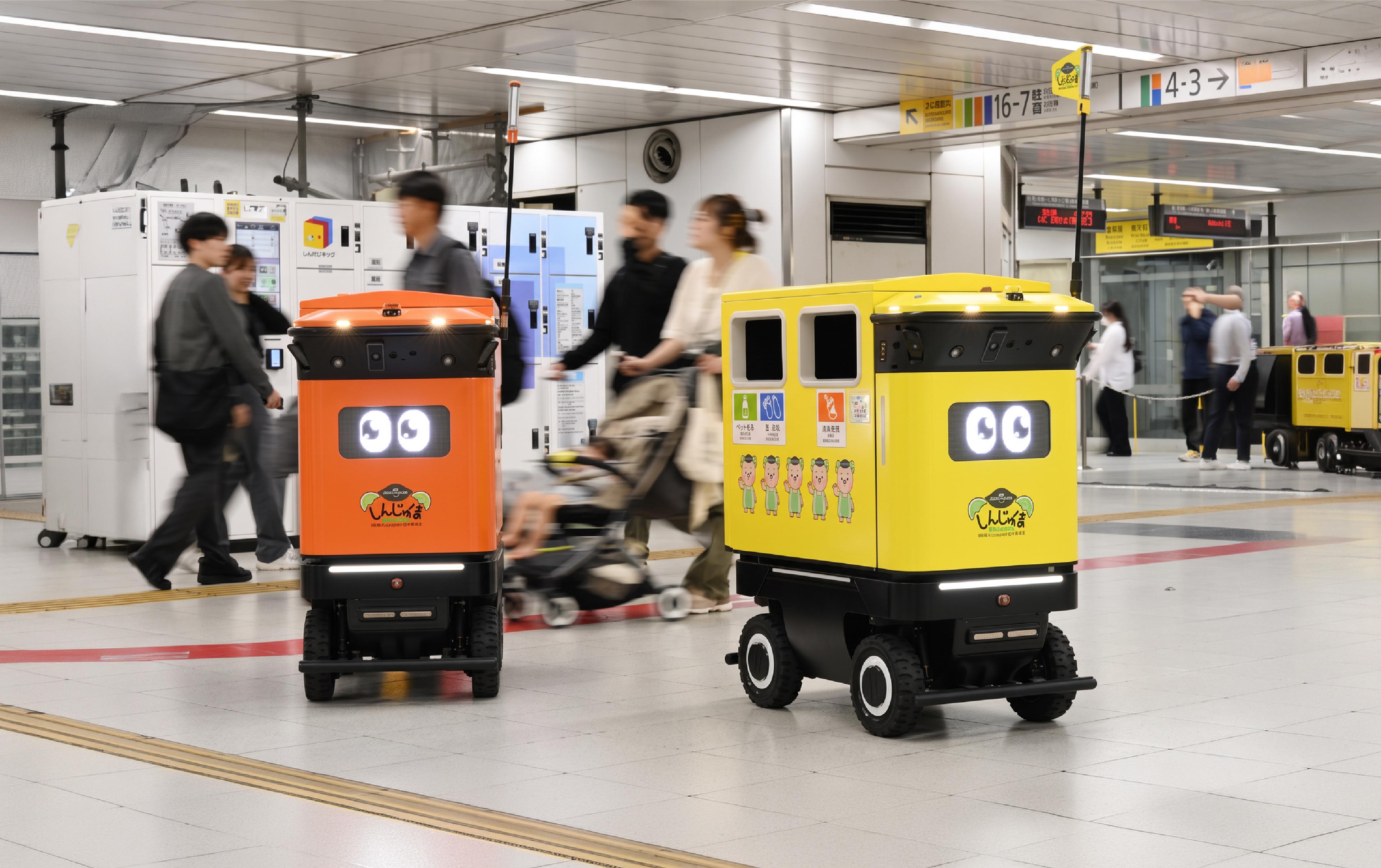 Two autonomous mobile rubbish-collection robots move through a busy Tokyo railway station concourse, with commuters walking and cycling around them.