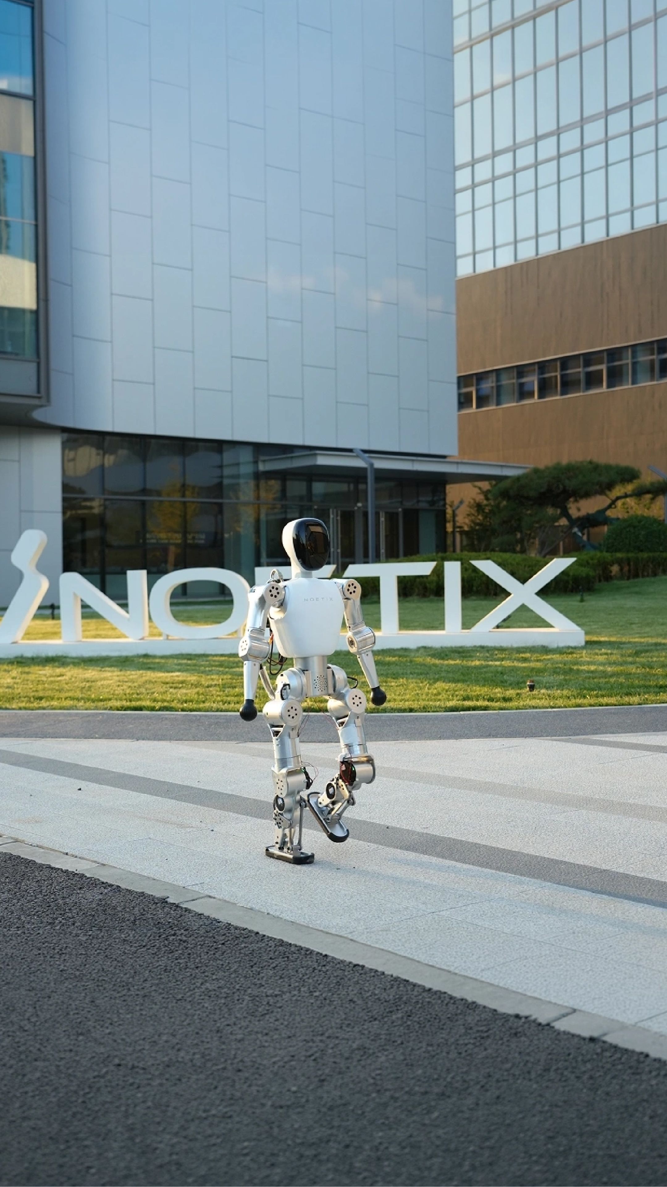 A white Noetix humanoid robot walks outdoors in front of the company's large logo and a modern office building.