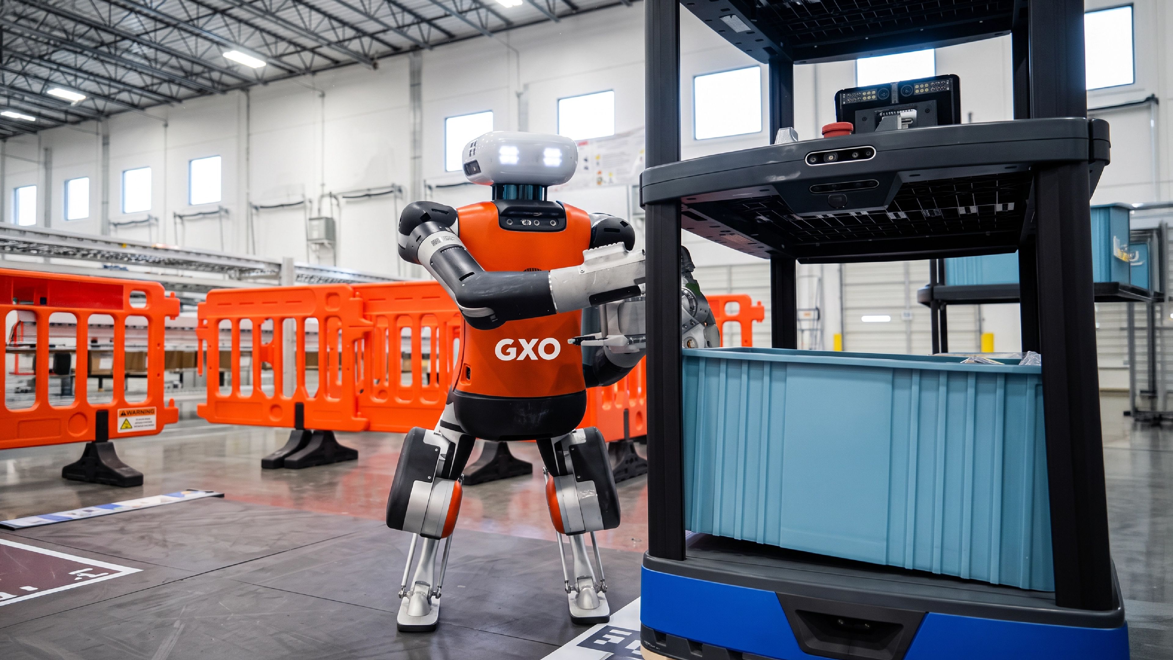 Agility Robotics' Digit humanoid robot, wearing GXO branding, handles a storage tote at a warehouse facility.