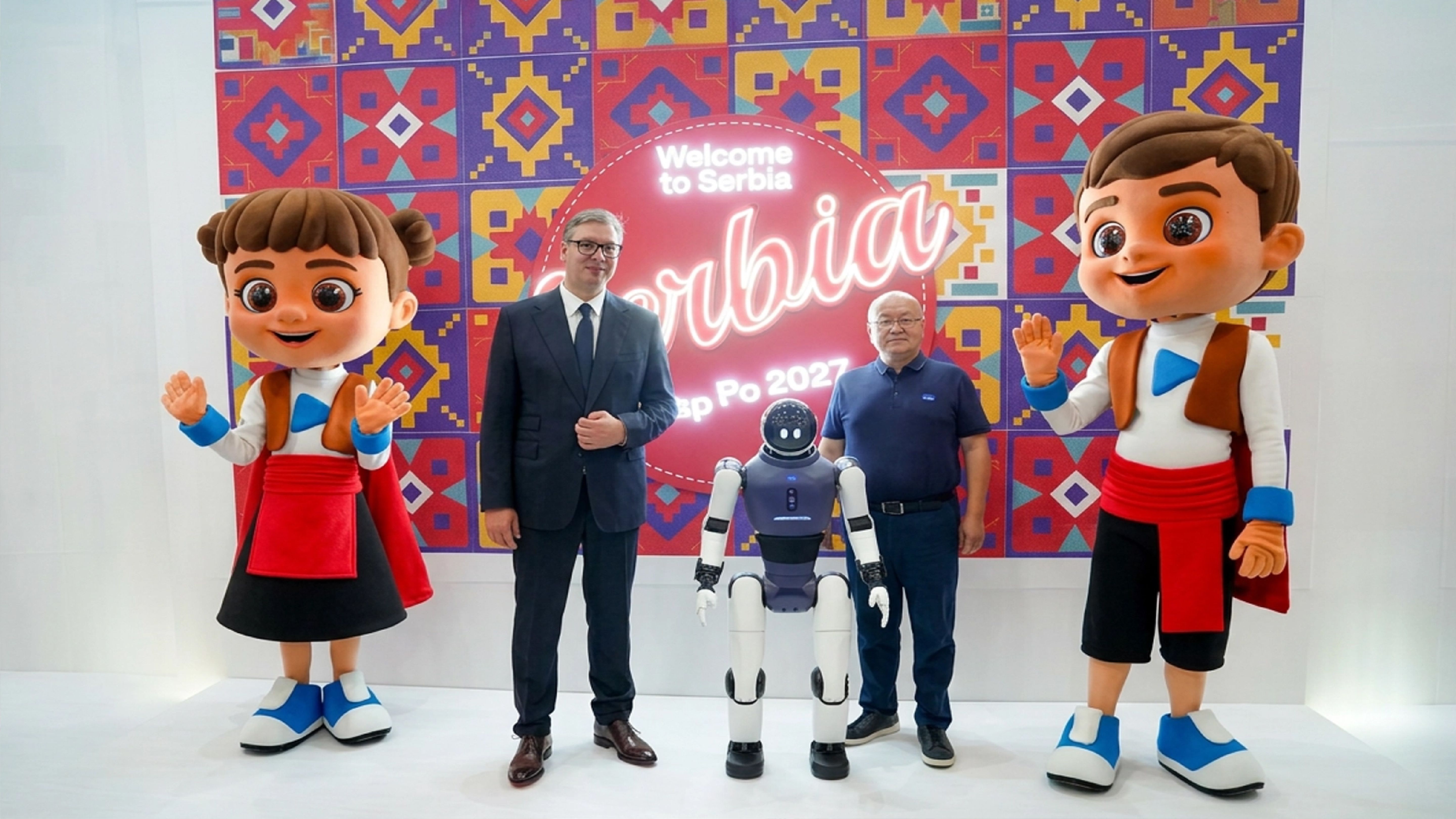 Humanoid robot between two men and two large mascots in front of a colourful “Welcome to Serbia” Expo 2027 backdrop.
