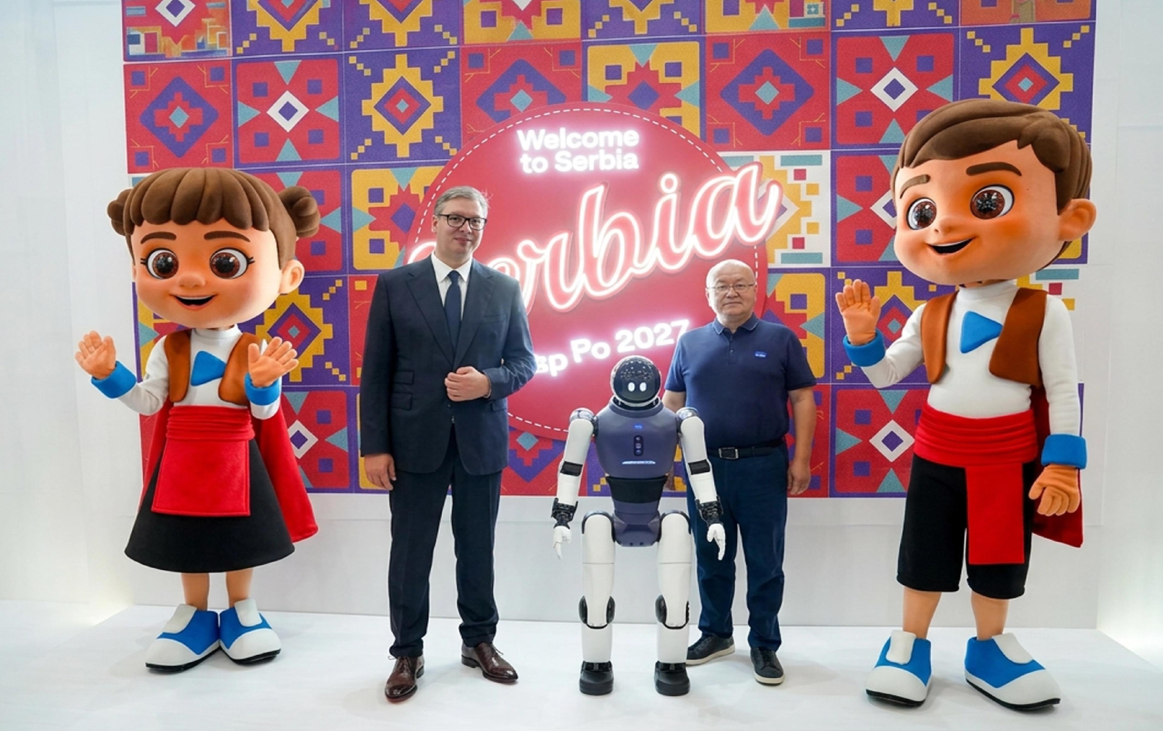 Humanoid robot between two men and two large mascots in front of a colourful “Welcome to Serbia” Expo 2027 backdrop.