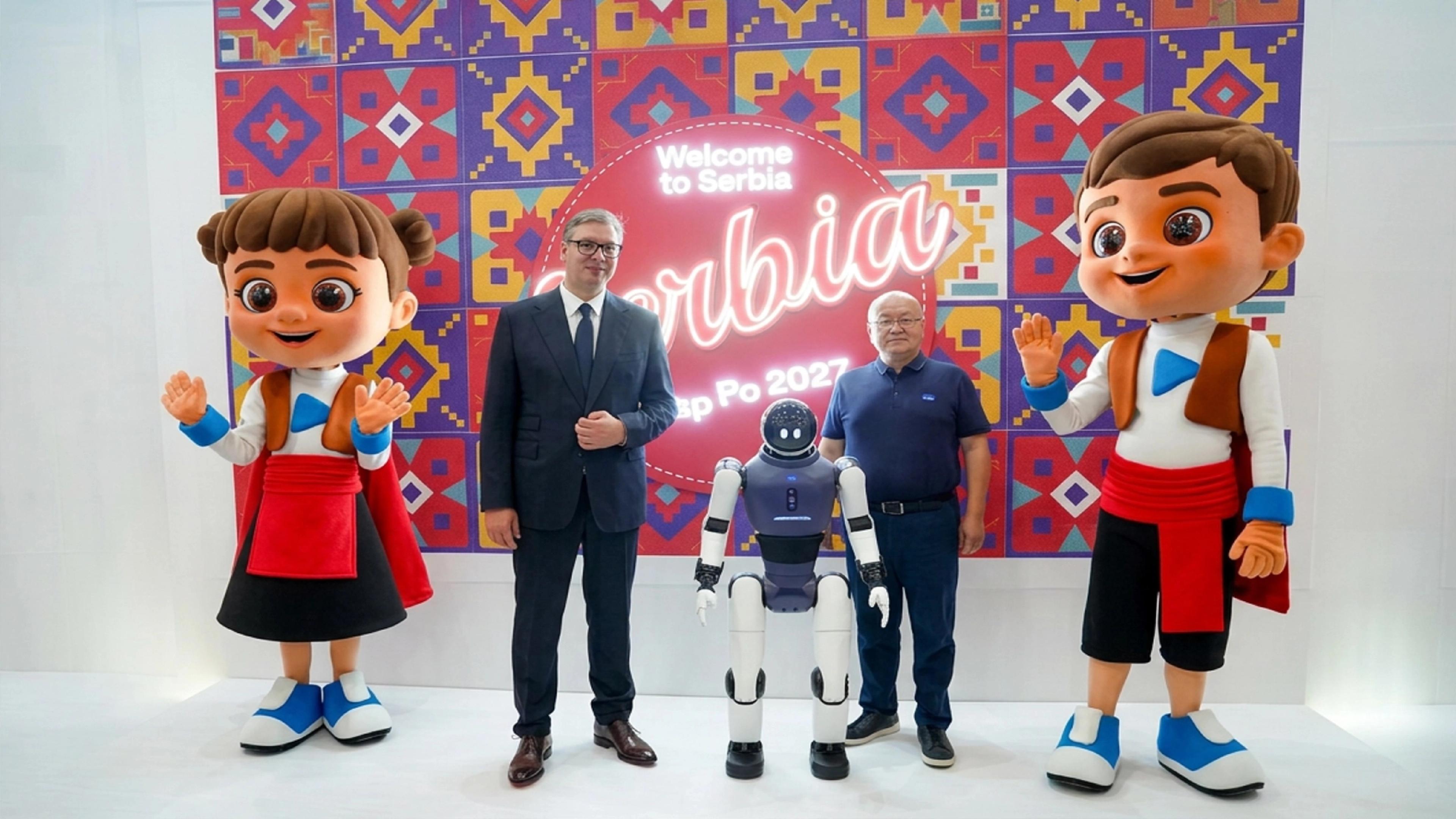 Humanoid robot between two men and two large mascots in front of a colourful “Welcome to Serbia” Expo 2027 backdrop.