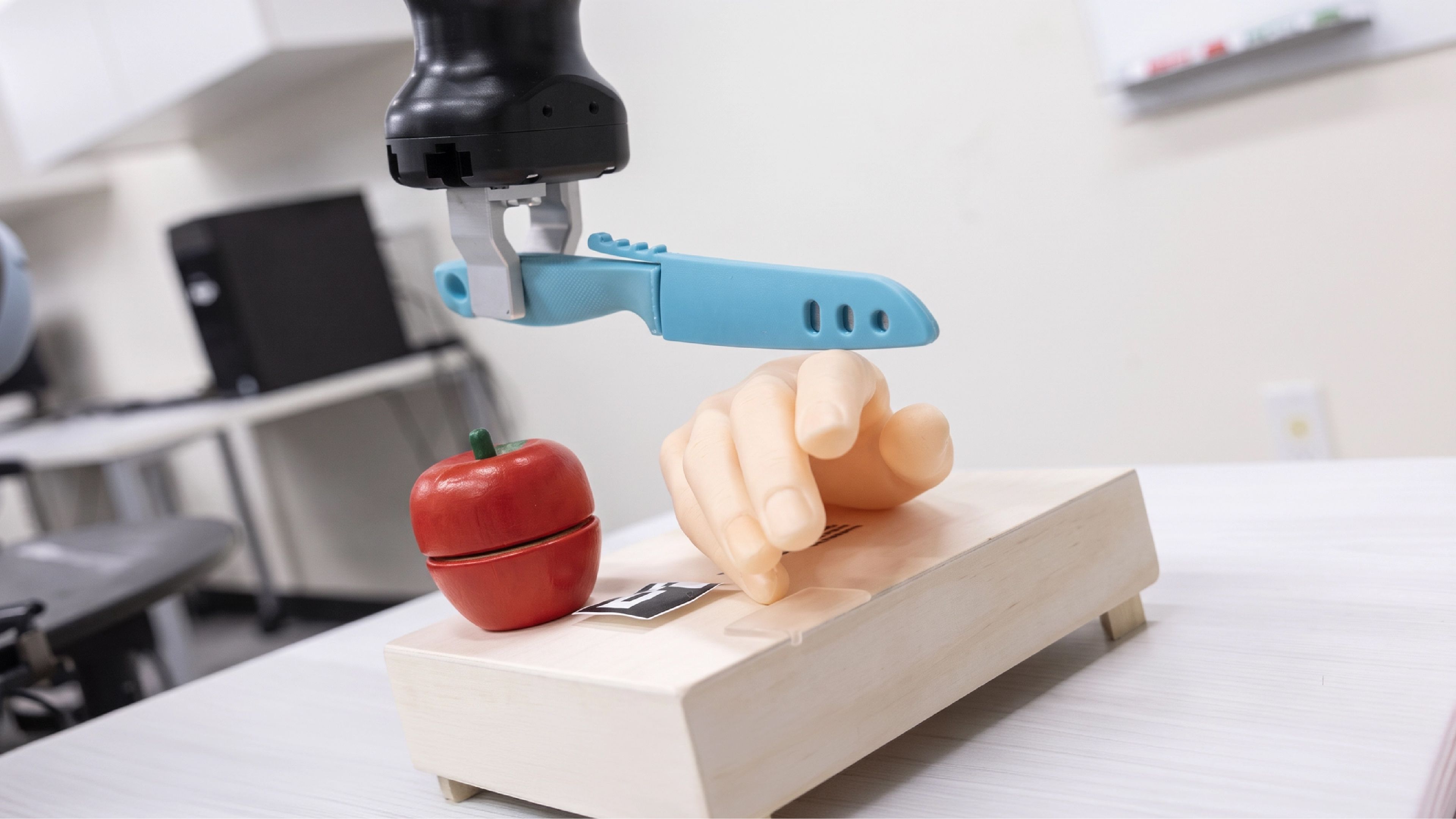 A robotic arm holds a plastic knife above a prosthetic hand and an apple in a lab setting.
