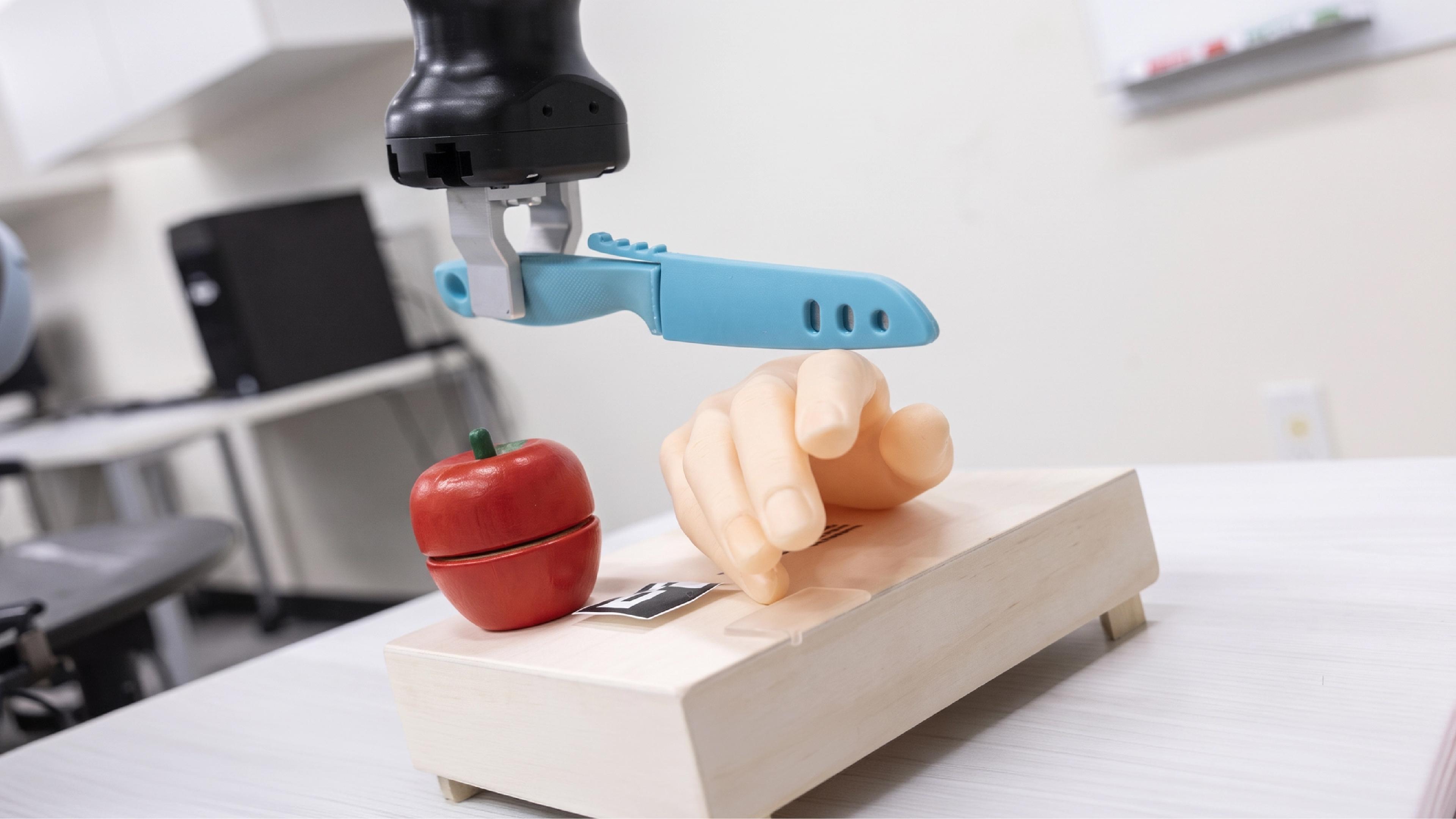 A robotic arm holds a plastic knife above a prosthetic hand and an apple in a lab setting.